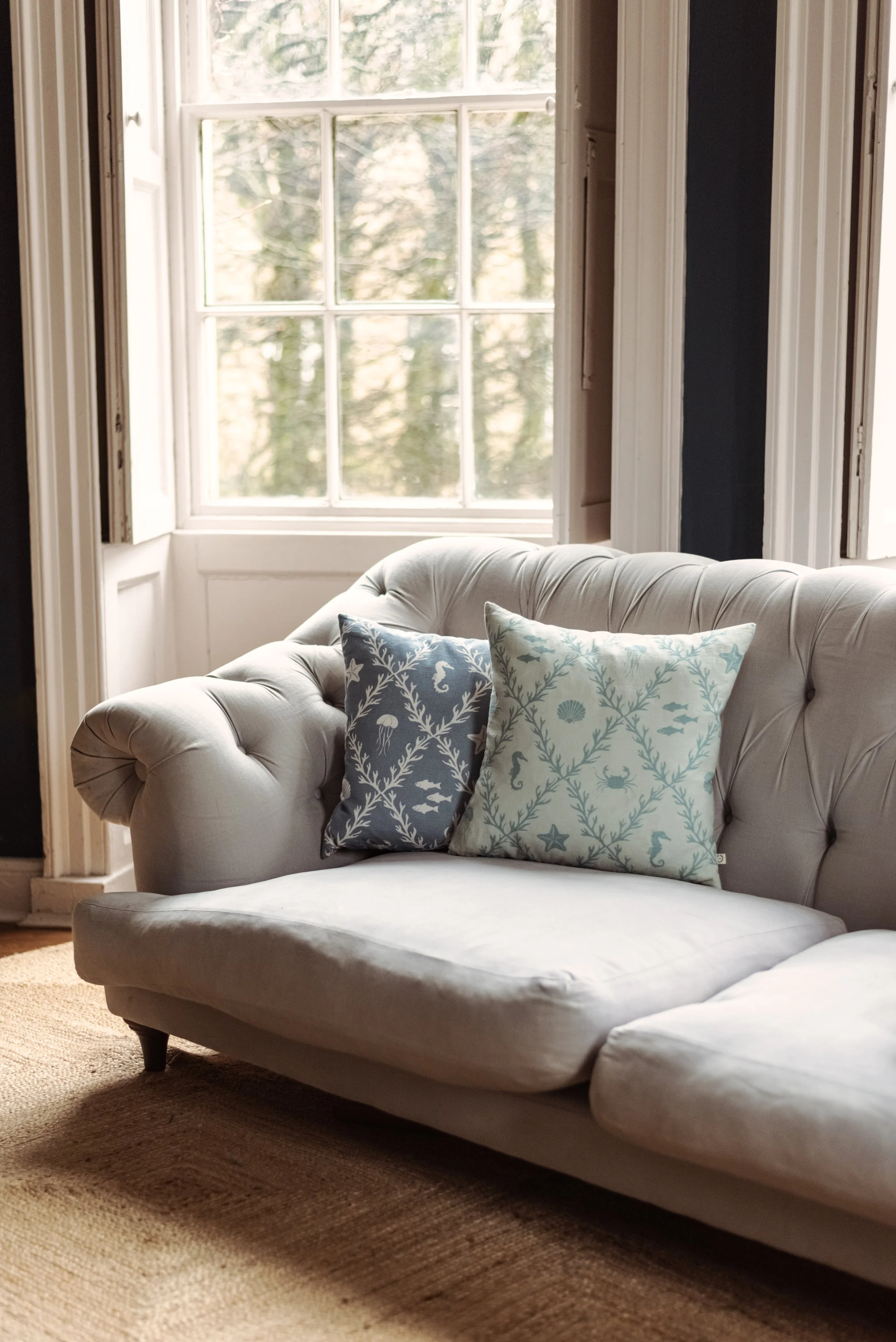 A beige tufted sofa with two patterned throw pillows, placed in front of a large window in a living room.