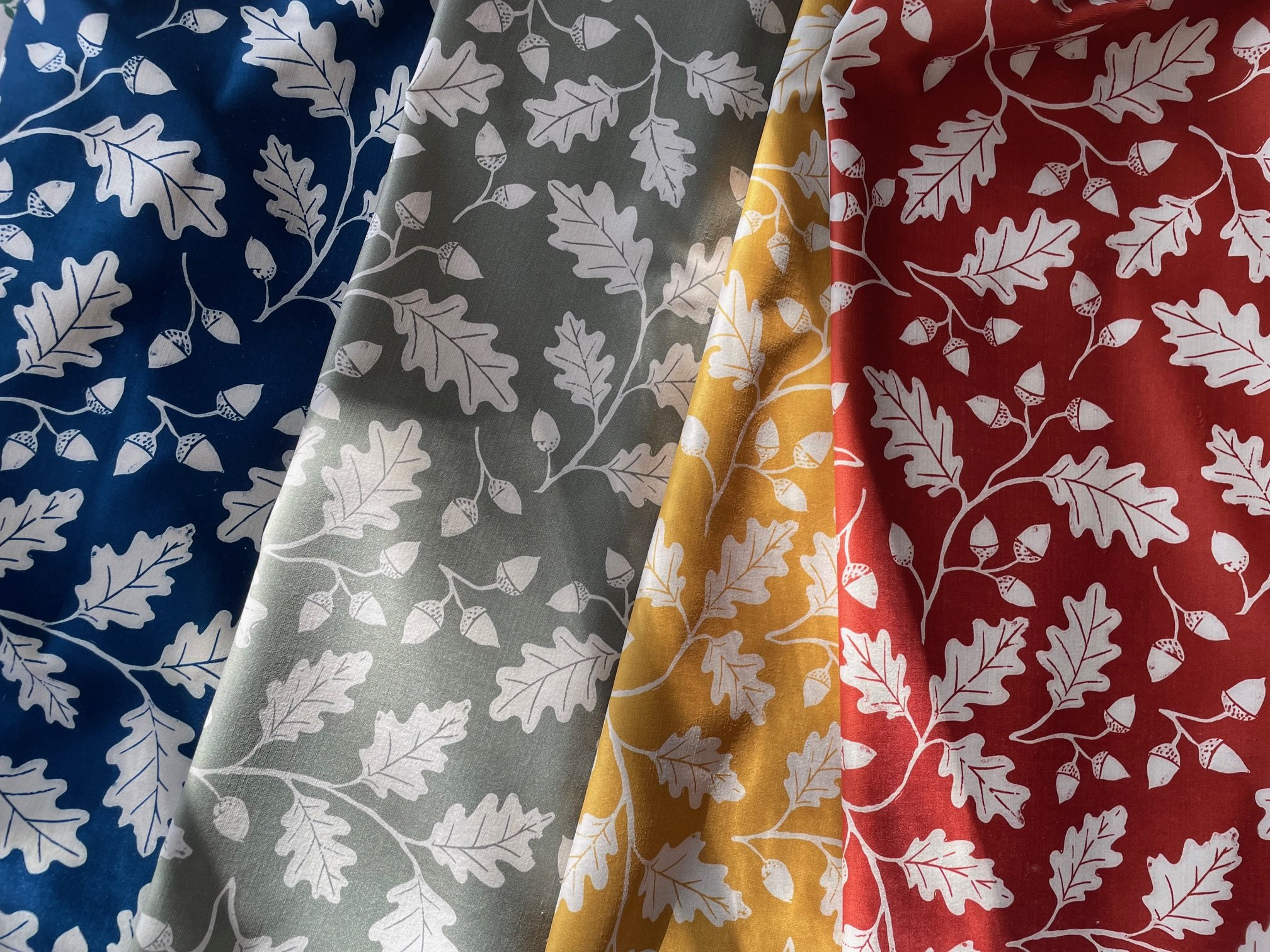 Four pieces of fabric with oak leaf and acorn patterns in blue, grey, yellow, and red colors, arranged diagonally.