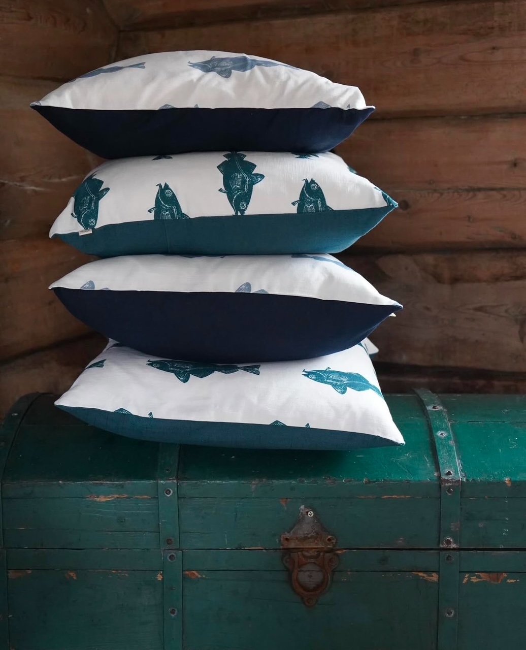 Four pillows with a blue and white fish pattern stacked on top of a green vintage trunk, set against a wooden wall background.