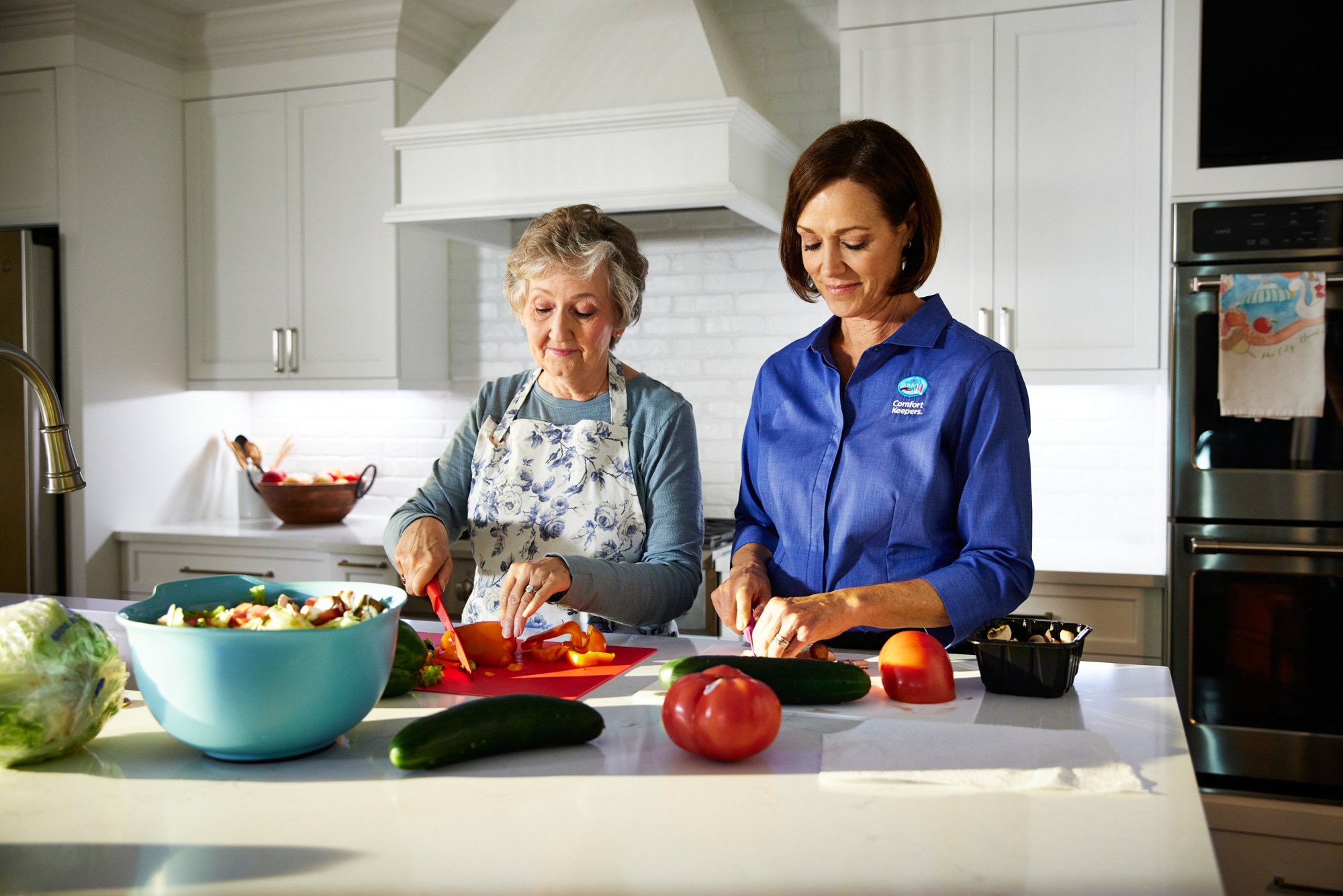 Two women preparing food in a modern kitchen, with vegetables and a blue mixing bowl on the counter.