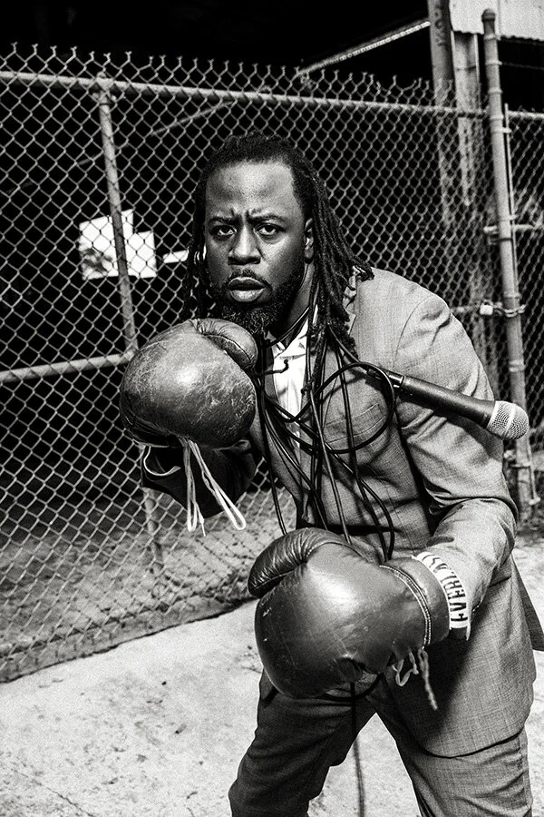 A man in a suit wearing boxing gloves is posing with a serious expression in front of a chain-link fence.