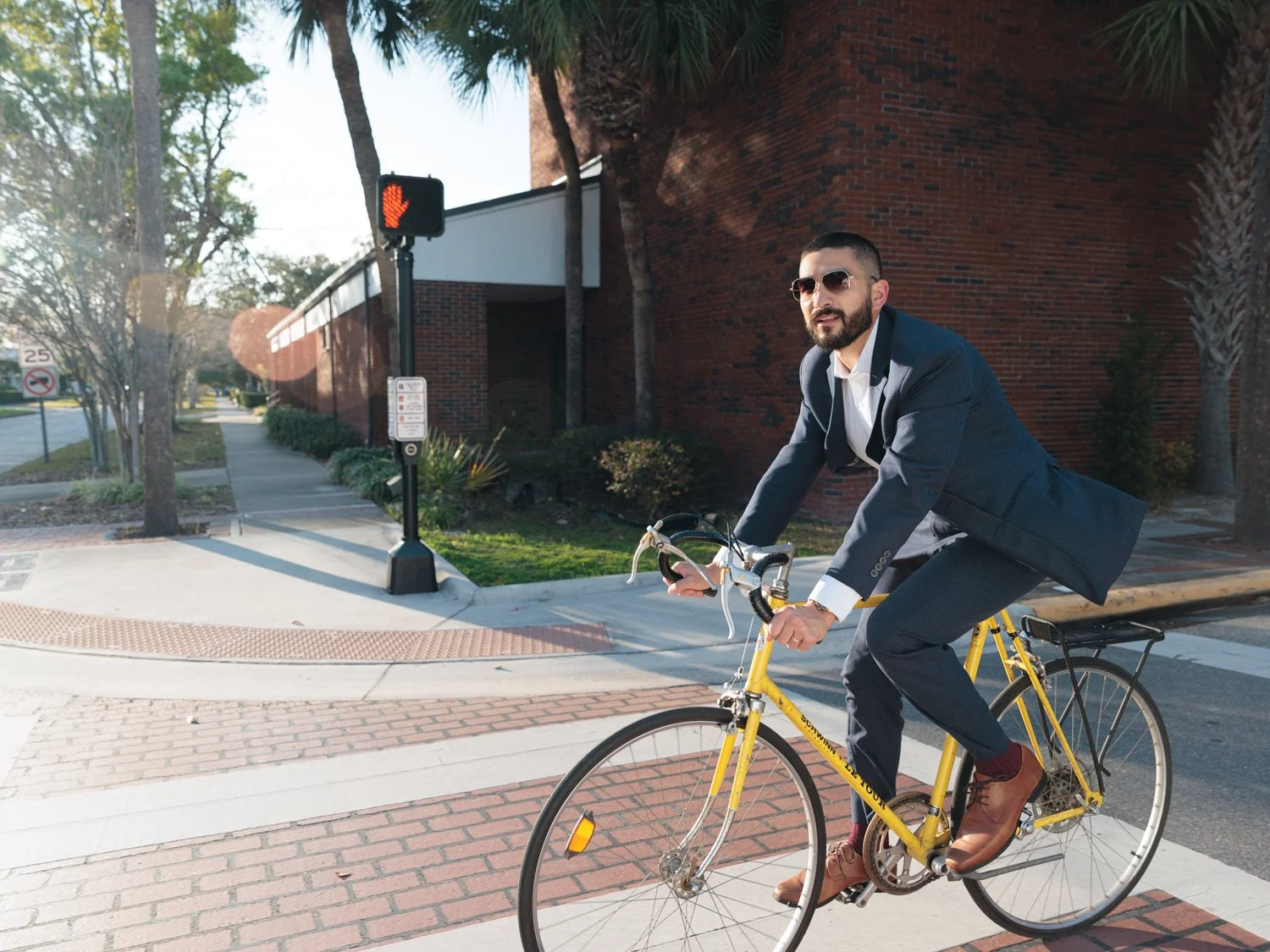 A man in a suit riding a yellow bicycle across a crosswalk in an urban area with trees and a brick building in the background.