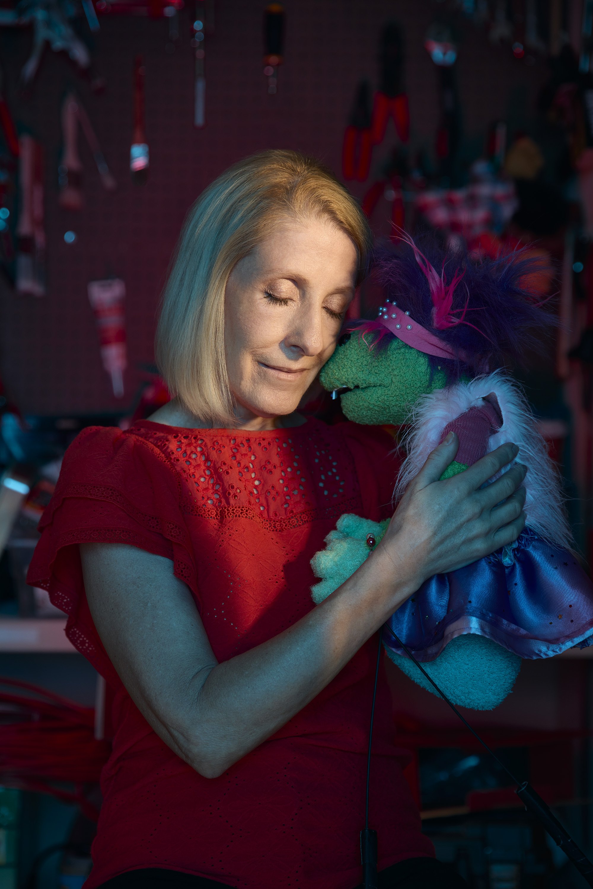 A woman with blonde hair and closed eyes embraces a colorful puppet with pink feathers and a green face, in a workshop setting with tools and ornaments hanging in the background.