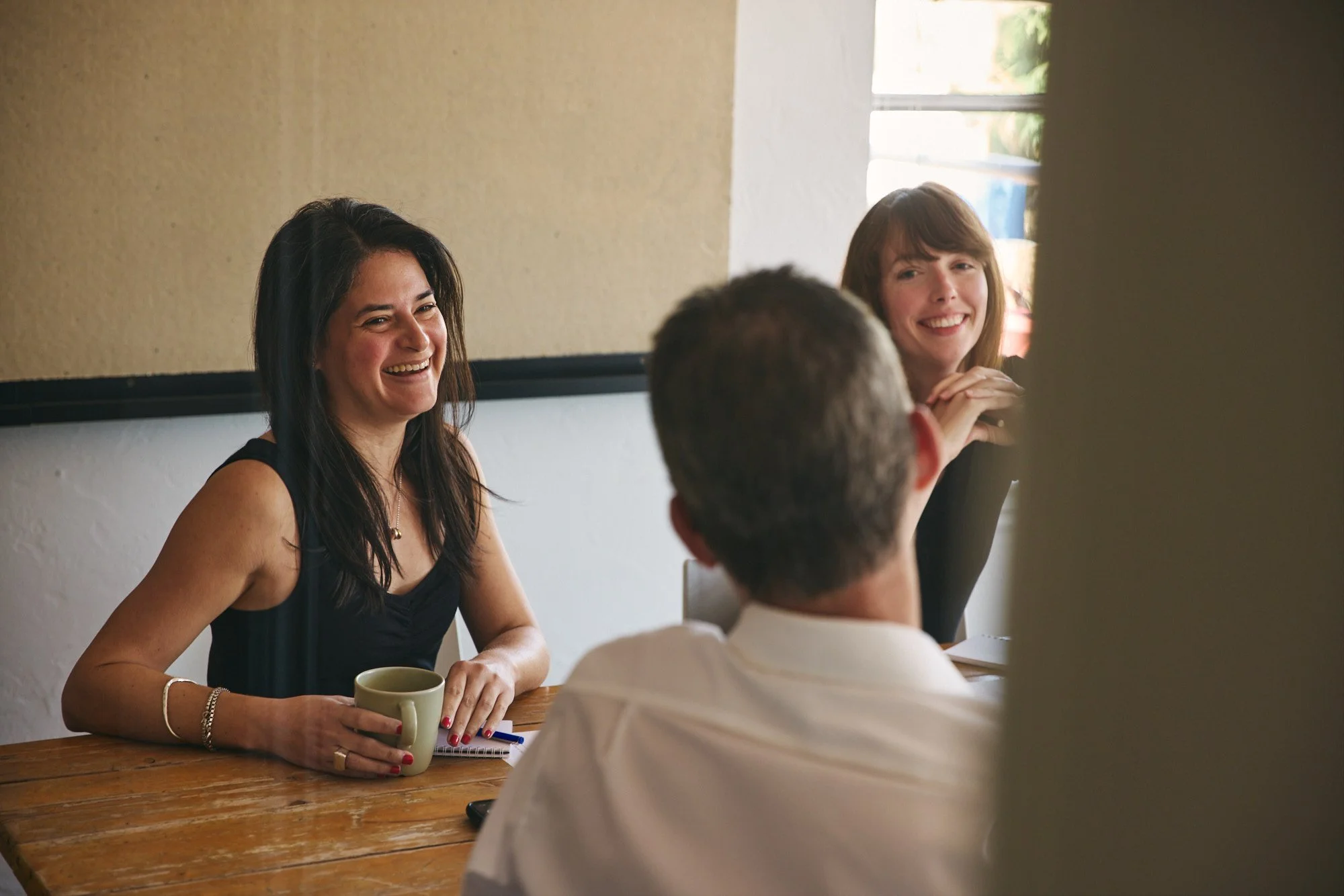 Three people are sitting at a wooden table in a room with a beige wall. Two women are smiling and talking, one with long dark hair wearing a black sleeveless top, and the other with light brown hair wearing a dark blazer. A man with short gray hair, seen from the back, is also at the table, holding a mug, engaged in conversation.