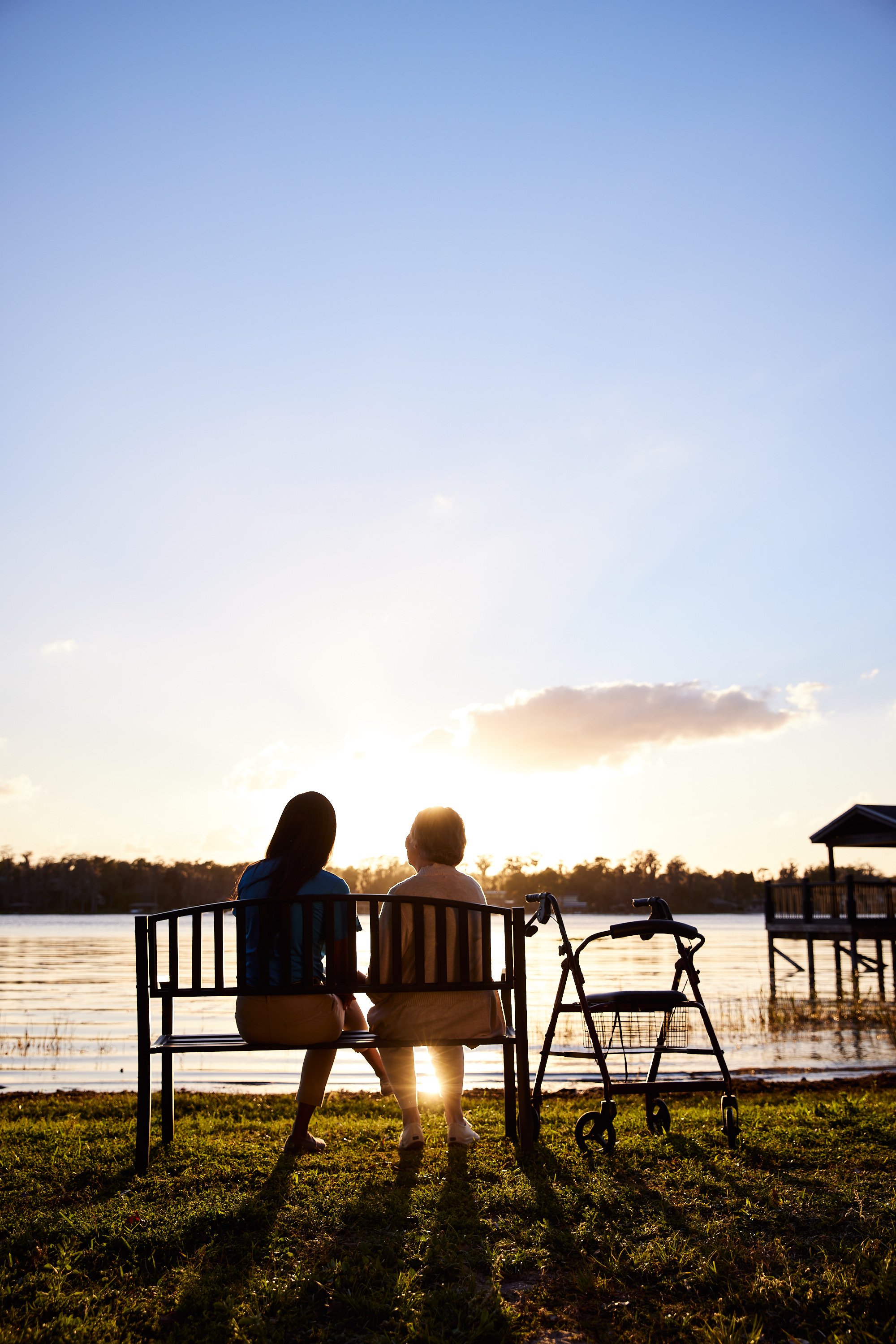 Two people sitting on a bench by a lake during sunset, with a walker nearby.
