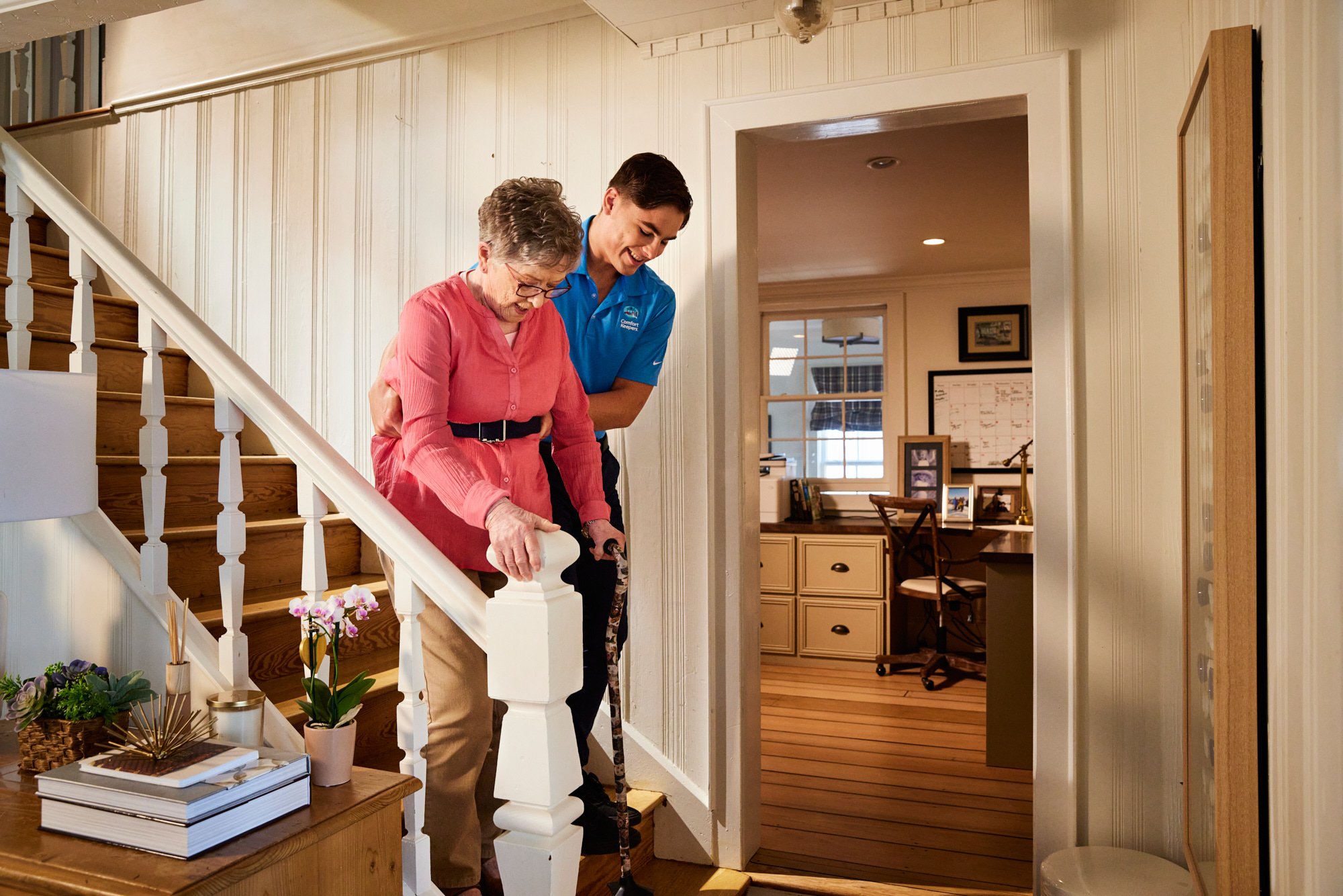 Elderly woman being assisted by a caregiver while walking down stairs inside a home.