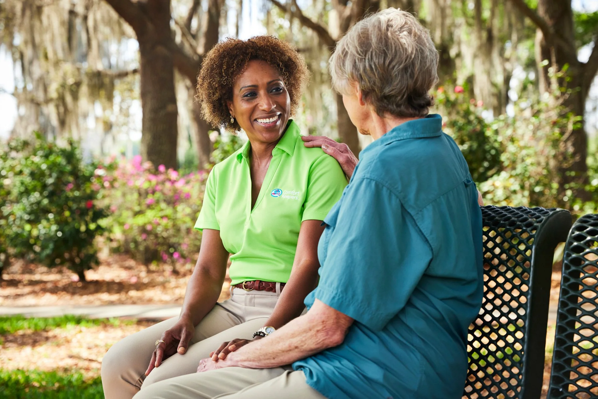 Two women sitting on a park bench, one in a green shirt labeled "Comfort Keepers," the other in a blue shirt, engaged in a conversation outdoors.