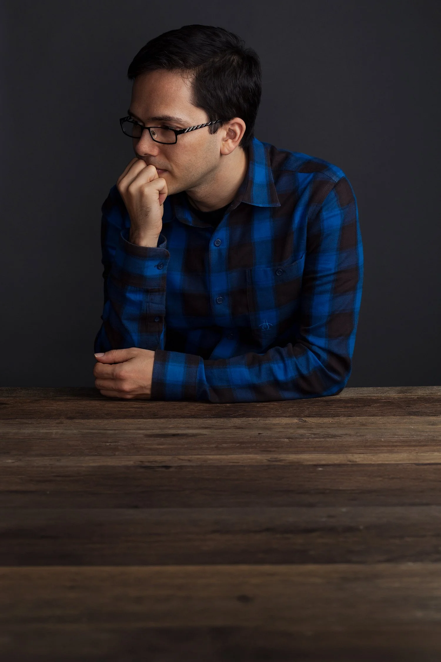 A young man with dark hair, glasses, and a blue plaid shirt sitting at a wooden table against a dark background, resting his chin on his hand with a contemplative expression.