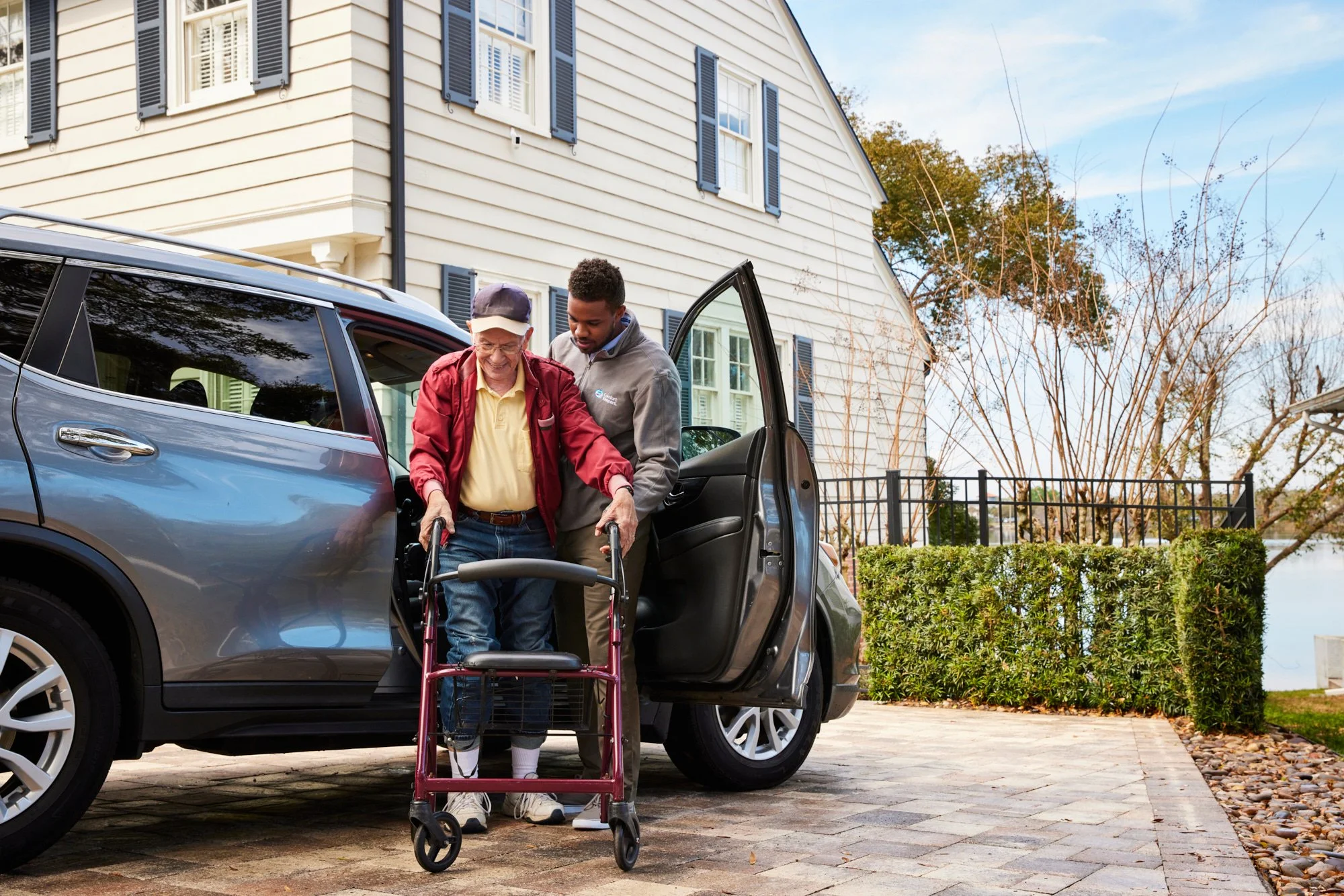 An elderly man using a walker exits a car with assistance from a younger man outside a house with shutters.