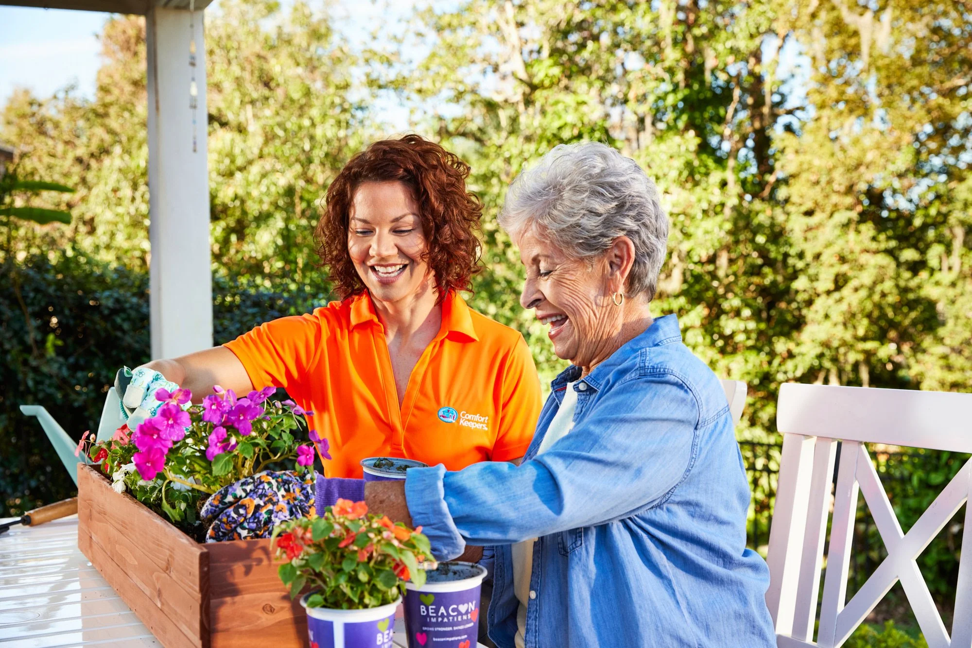 Two women gardening, one in an orange "Comfort Keepers" shirt, smiling and planting flowers in a wooden box on an outdoor patio.