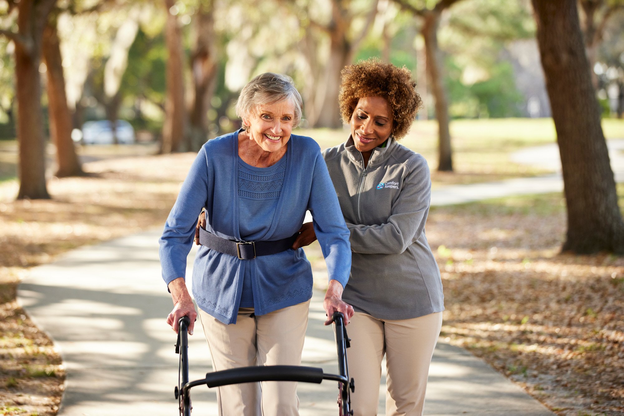 An elderly woman using a walker is assisted by a caregiver in a park setting.