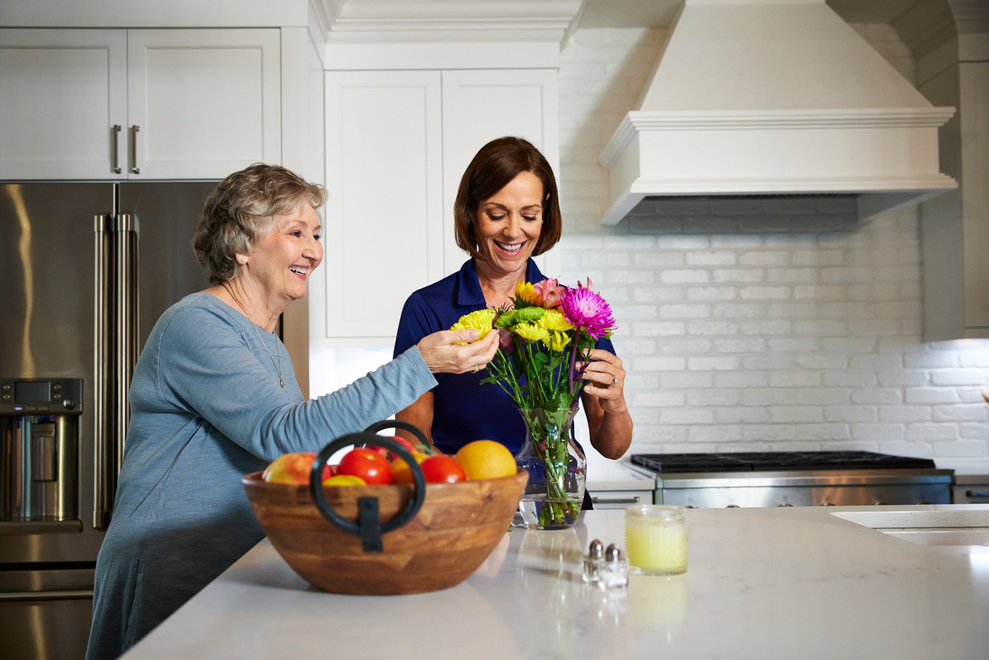 Two women arranging flowers in a kitchen with white cabinets and a wooden bowl of fruit on the counter.