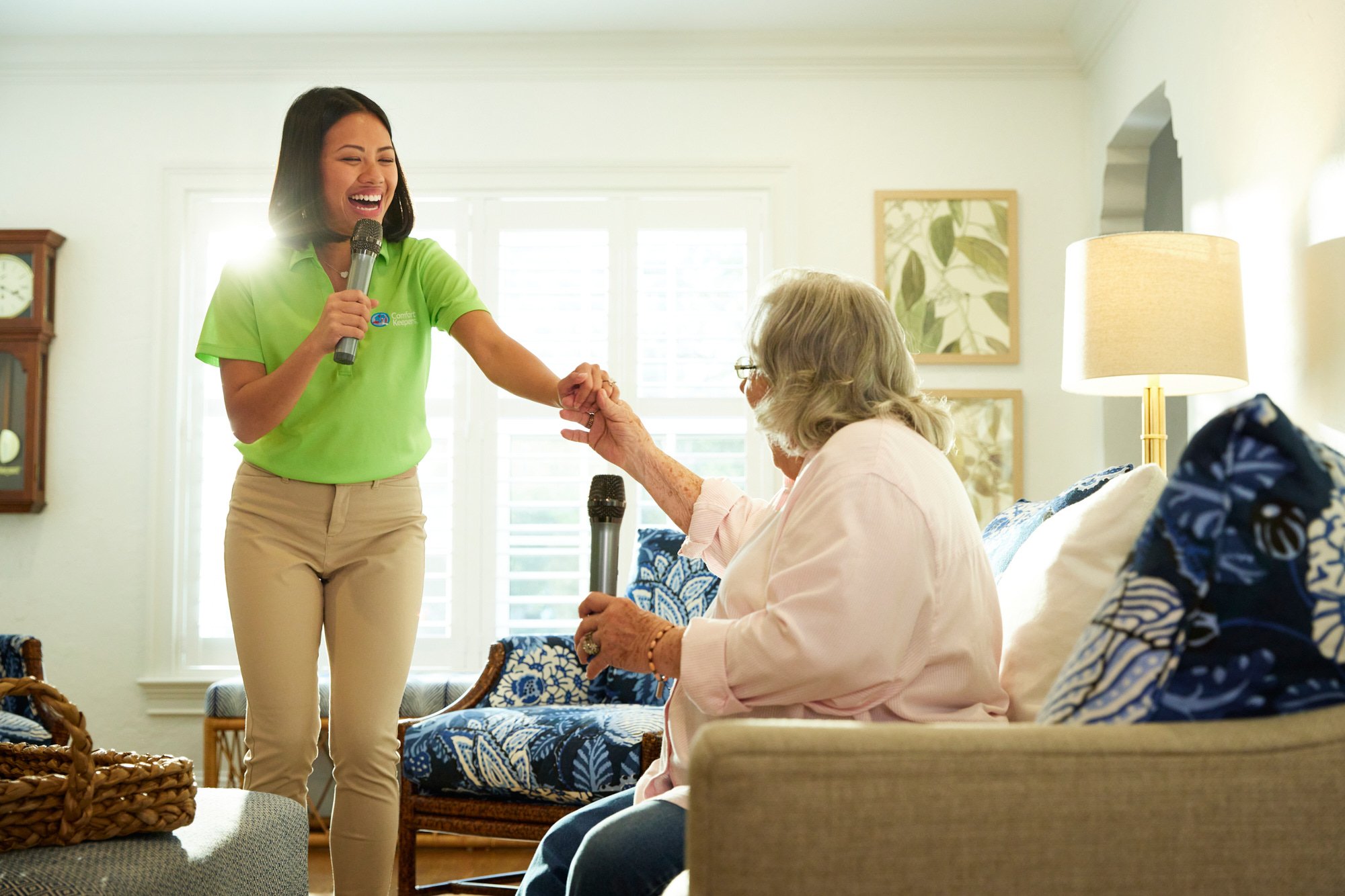 A caregiver engages in karaoke with a senior woman in a living room setting. The caregiver is smiling and holding a microphone while standing, and the senior woman is seated with another microphone. The room features patterned cushions and a clock on