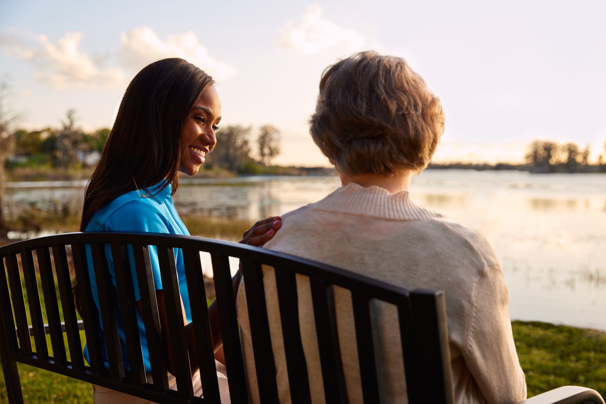 Two women sitting on a bench by a lake, one young woman smiling at an older woman, in a serene outdoor setting.