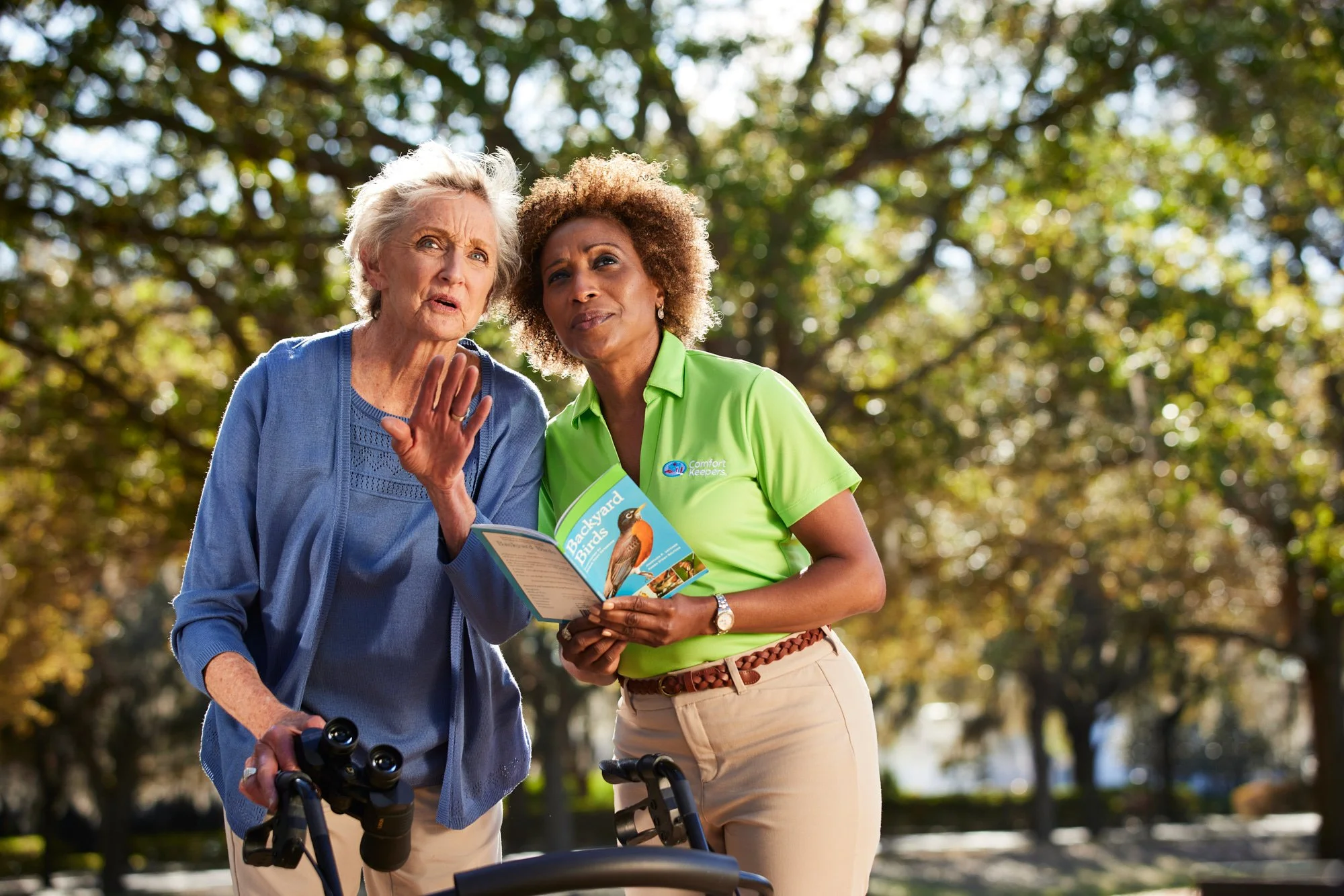 Elderly woman with binoculars and a caregiver reading a bird guide outdoors.