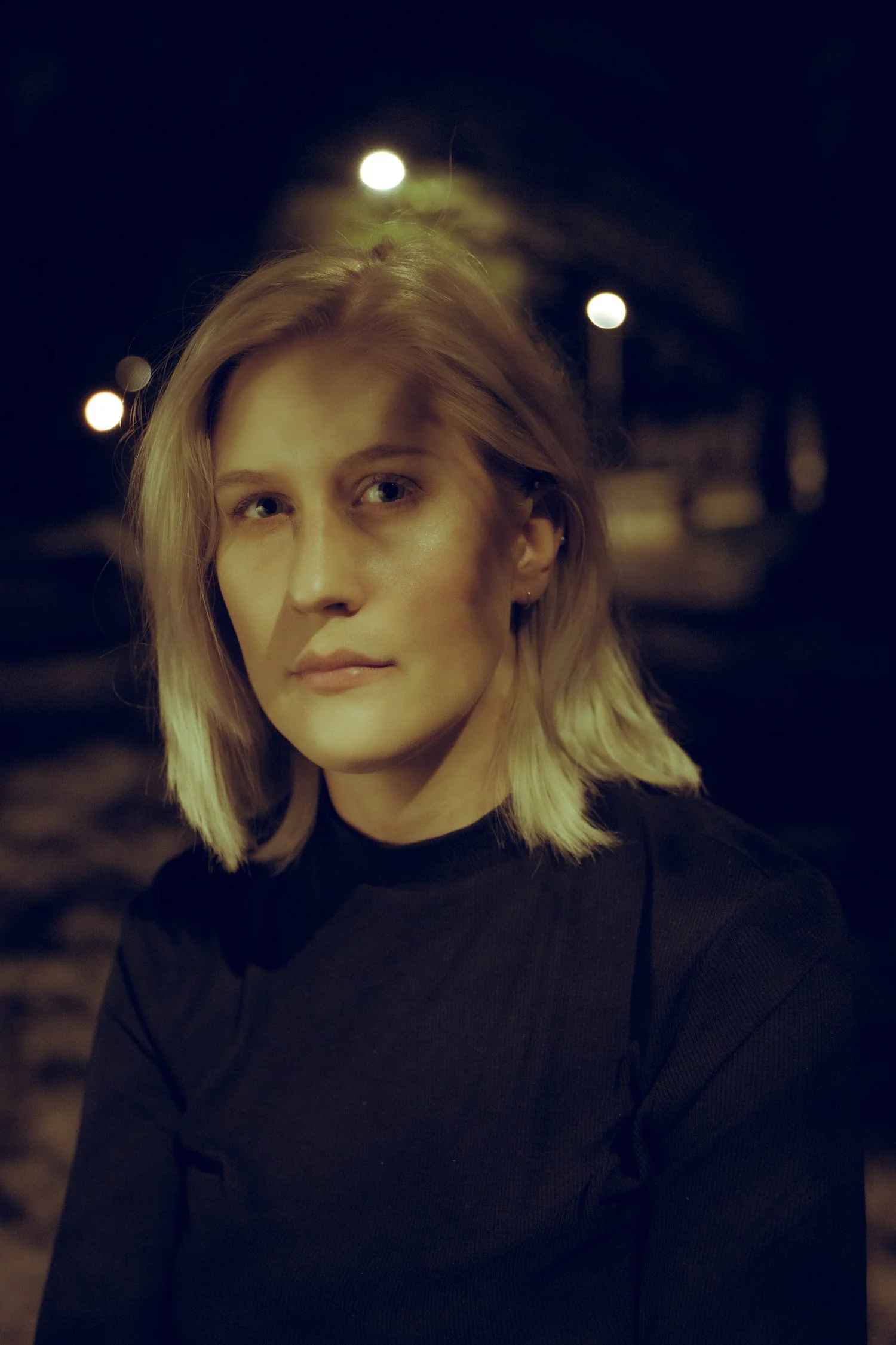 Close-up portrait of a blonde woman with short hair, wearing a black top, against a dark background with blurred streetlights at night.