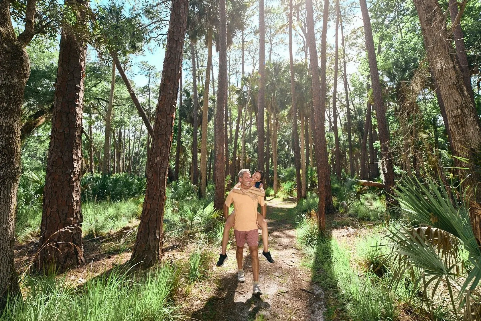 photo of man carrying girlfriend on back and laughing while on a hike in the woods