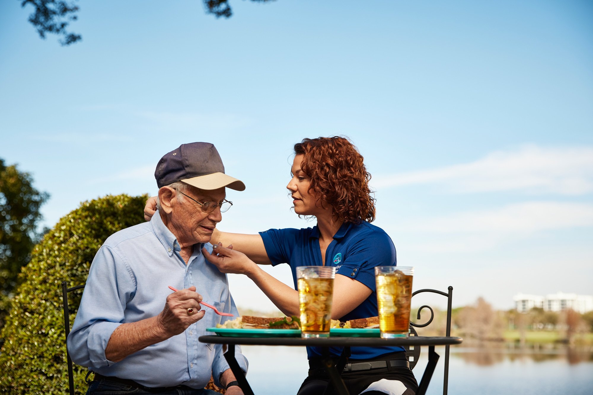 A senior man and a caregiver sitting outdoors at a table beside a lake, enjoying a meal with iced tea.