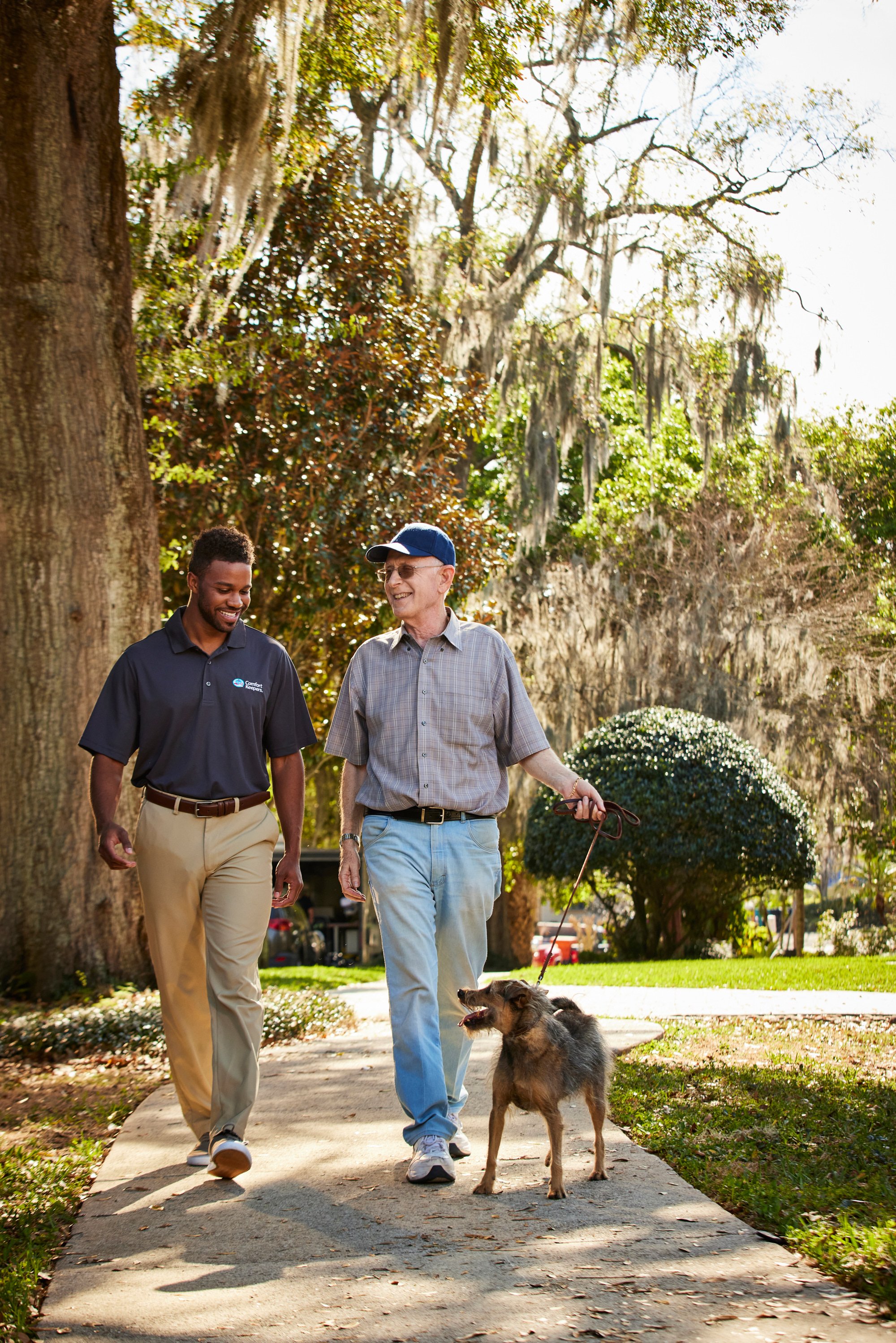 Two men walking on a sidewalk with a dog on a leash, surrounded by trees in a park setting.
