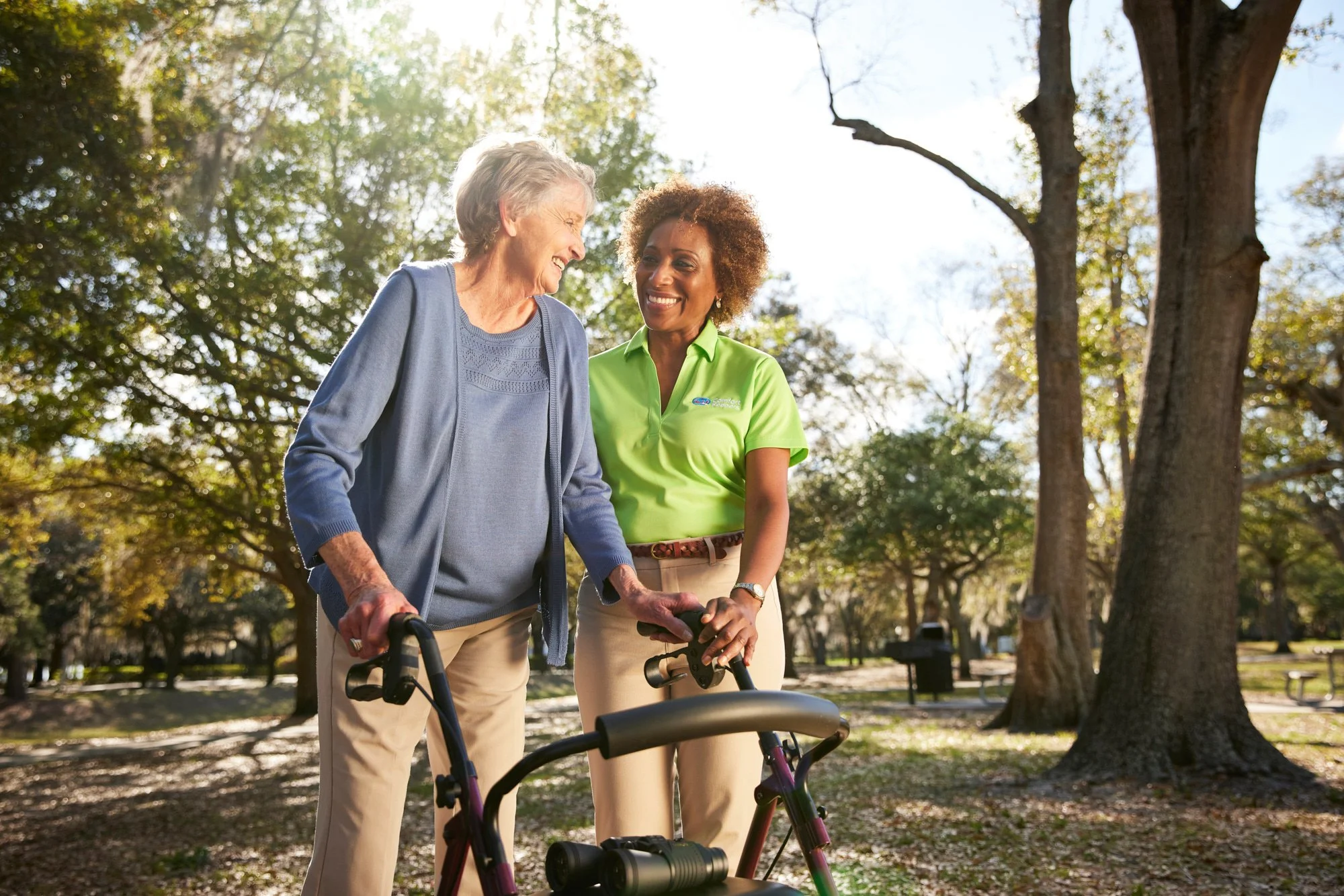 An elderly woman with a walker smiling and walking with a caregiver in a green shirt in a park.