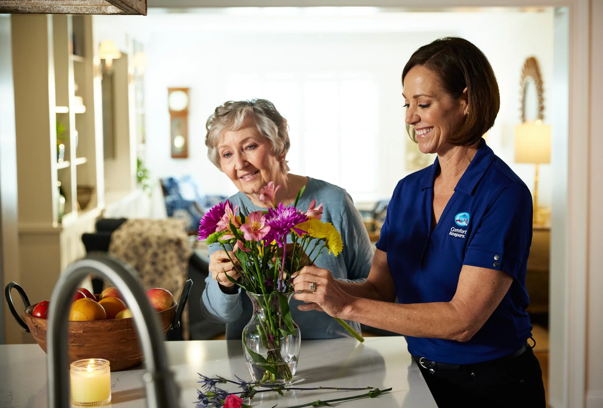 Two women arranging flowers in a vase on a kitchen counter with a bowl of fruit and a candle nearby.