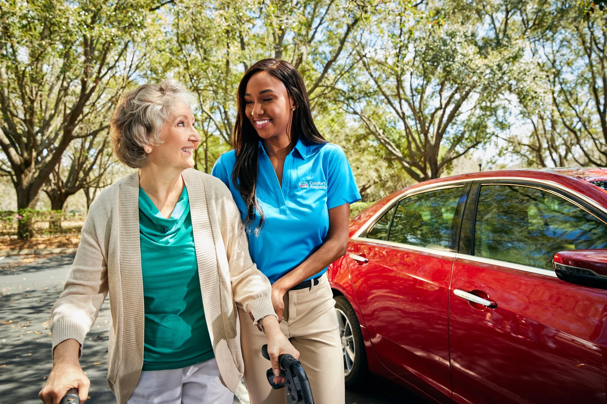 A caregiver in a blue shirt assists an elderly woman using a walker, next to a red car in a park with trees.