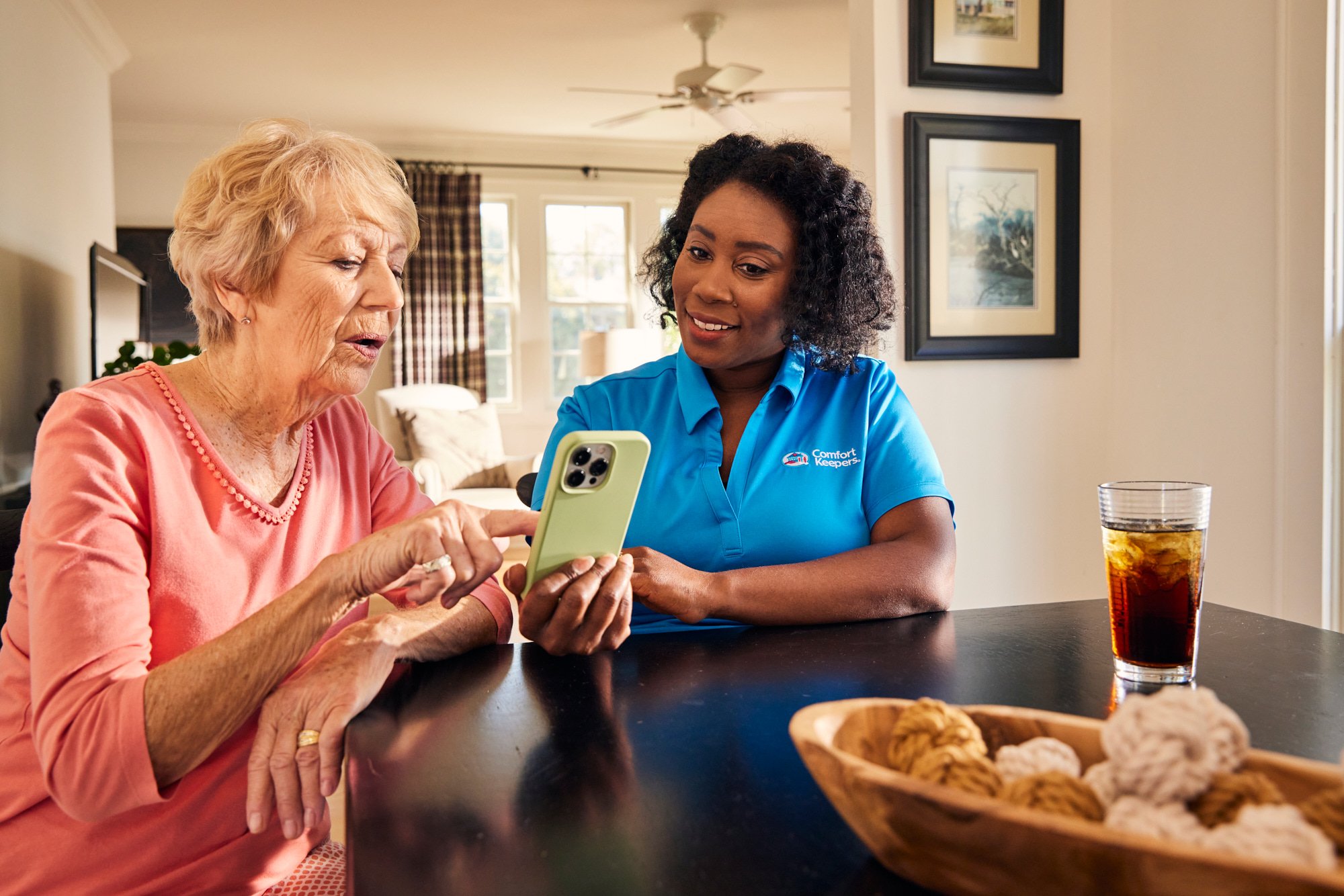 Elderly woman and caregiver using a smartphone at a table with cookies and drink in a living room.