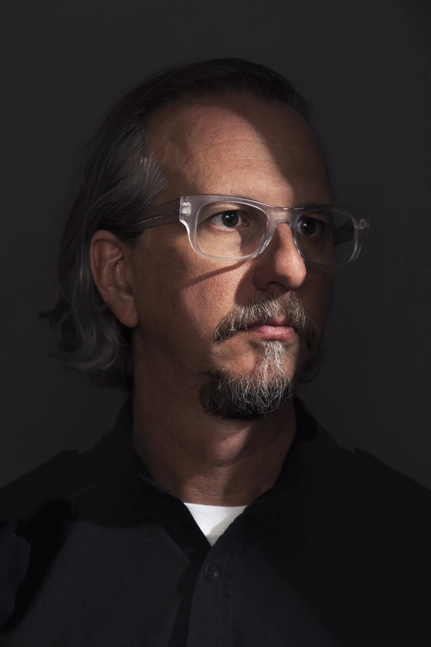 Close-up portrait of a middle-aged man with salt-and-pepper hair and beard, wearing clear safety glasses, against a dark background.