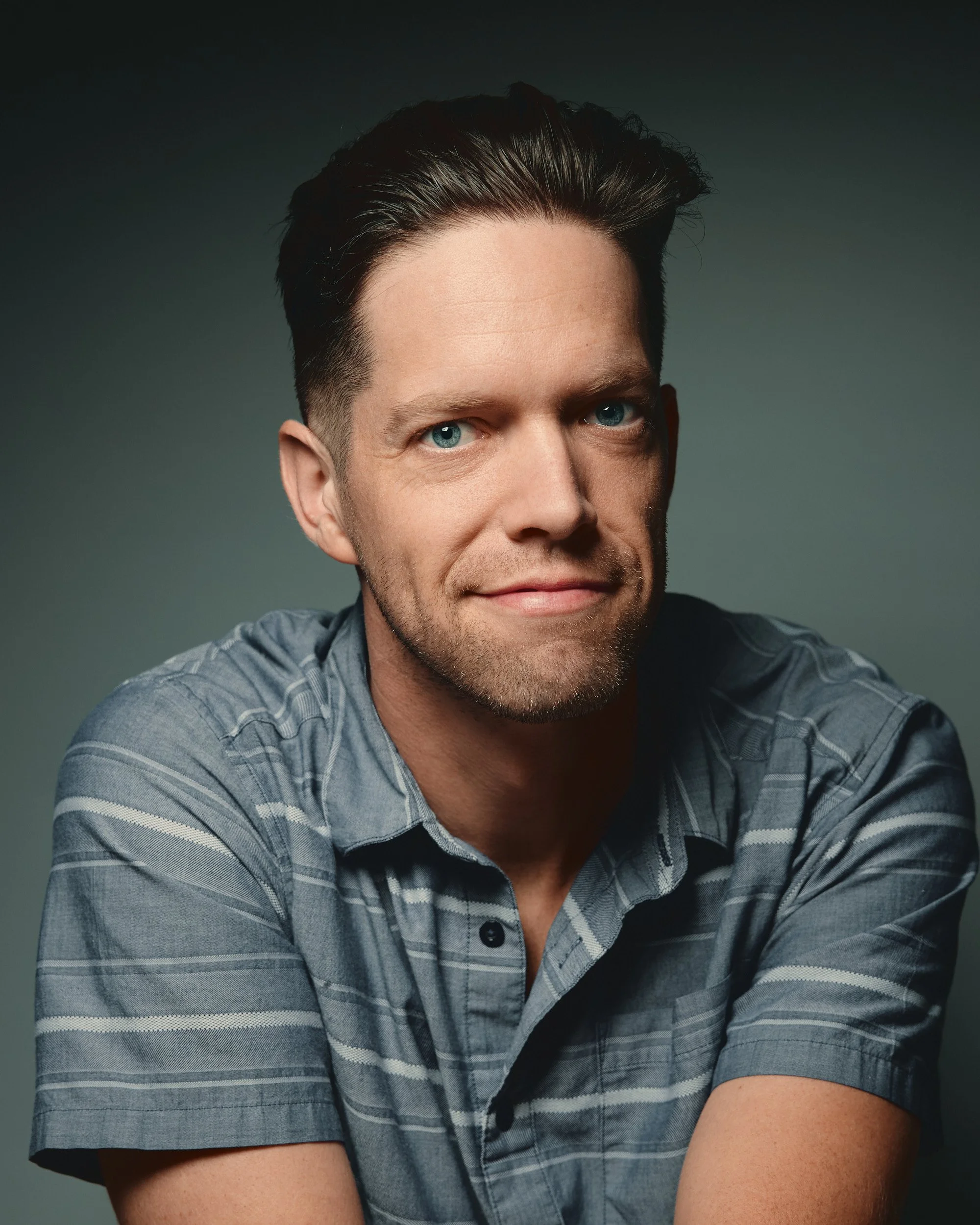 Half of a man’s face with blue eyes, dark hair styled upward, wearing a gray and black striped shirt, against a dark gray background.