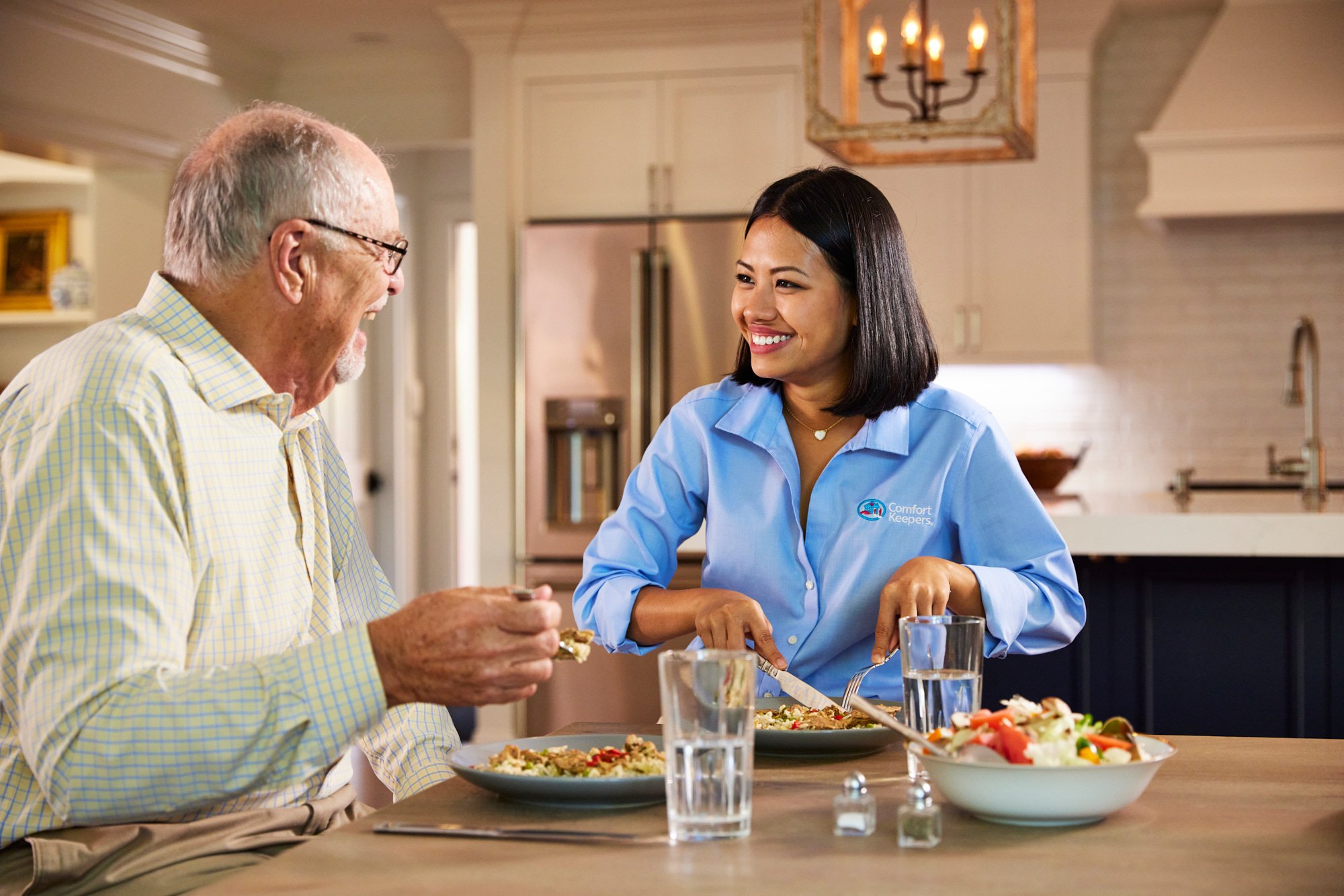 Elderly man and caregiver sharing a meal in a modern kitchen, smiling and conversing.