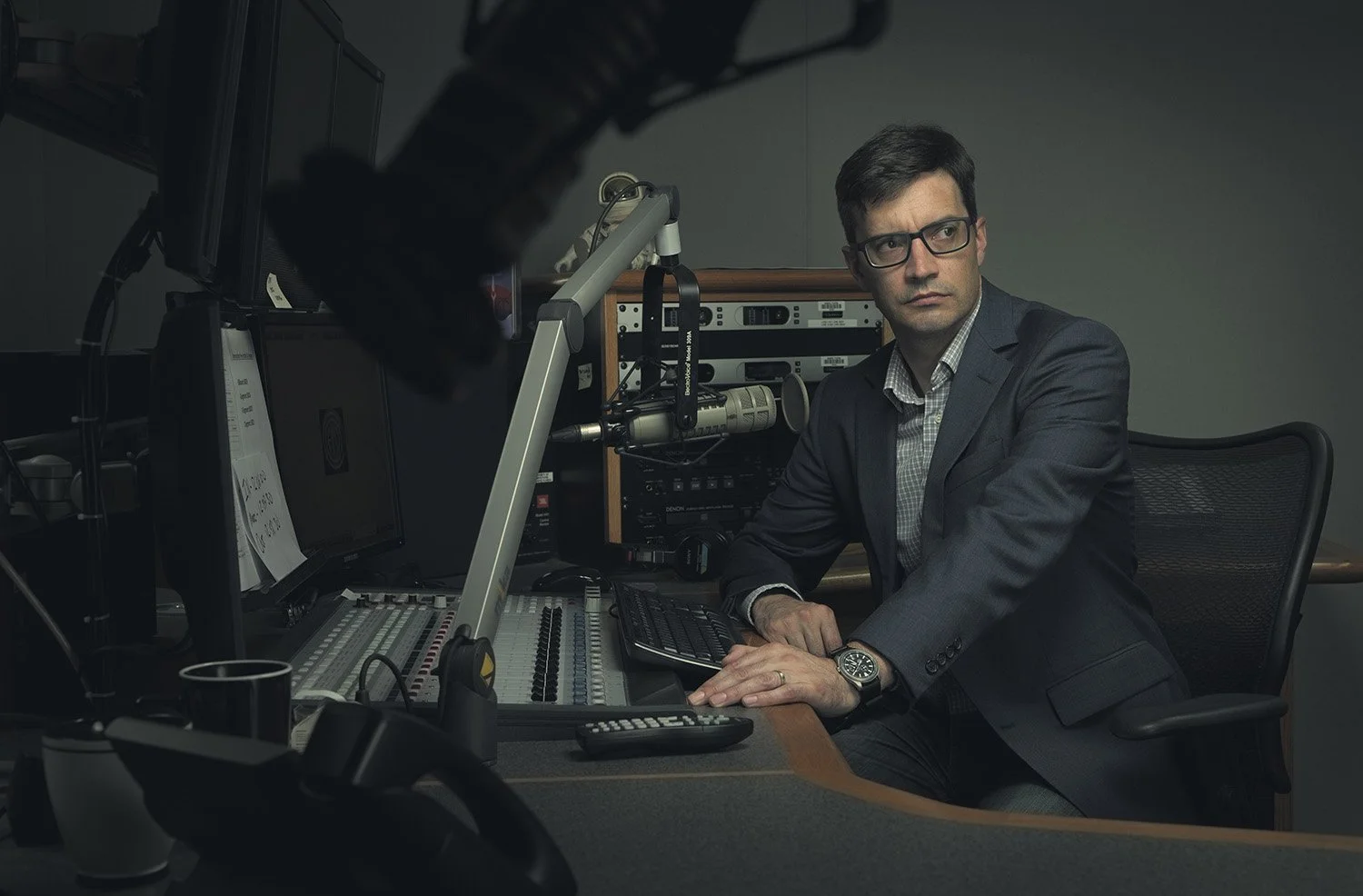 A man in glasses and a dark suit sitting at a radio broadcast station, with audio equipment, a microphone, and computer monitors in the background.