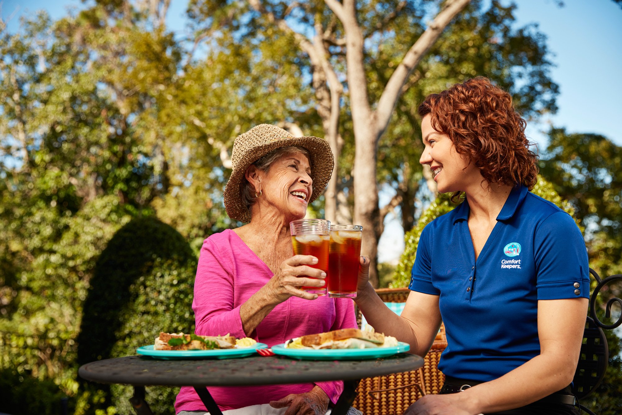 Two women clinking glasses of iced tea outdoors, one wearing a hat and pink shirt, the other in a blue uniform, with plates of food on a table.