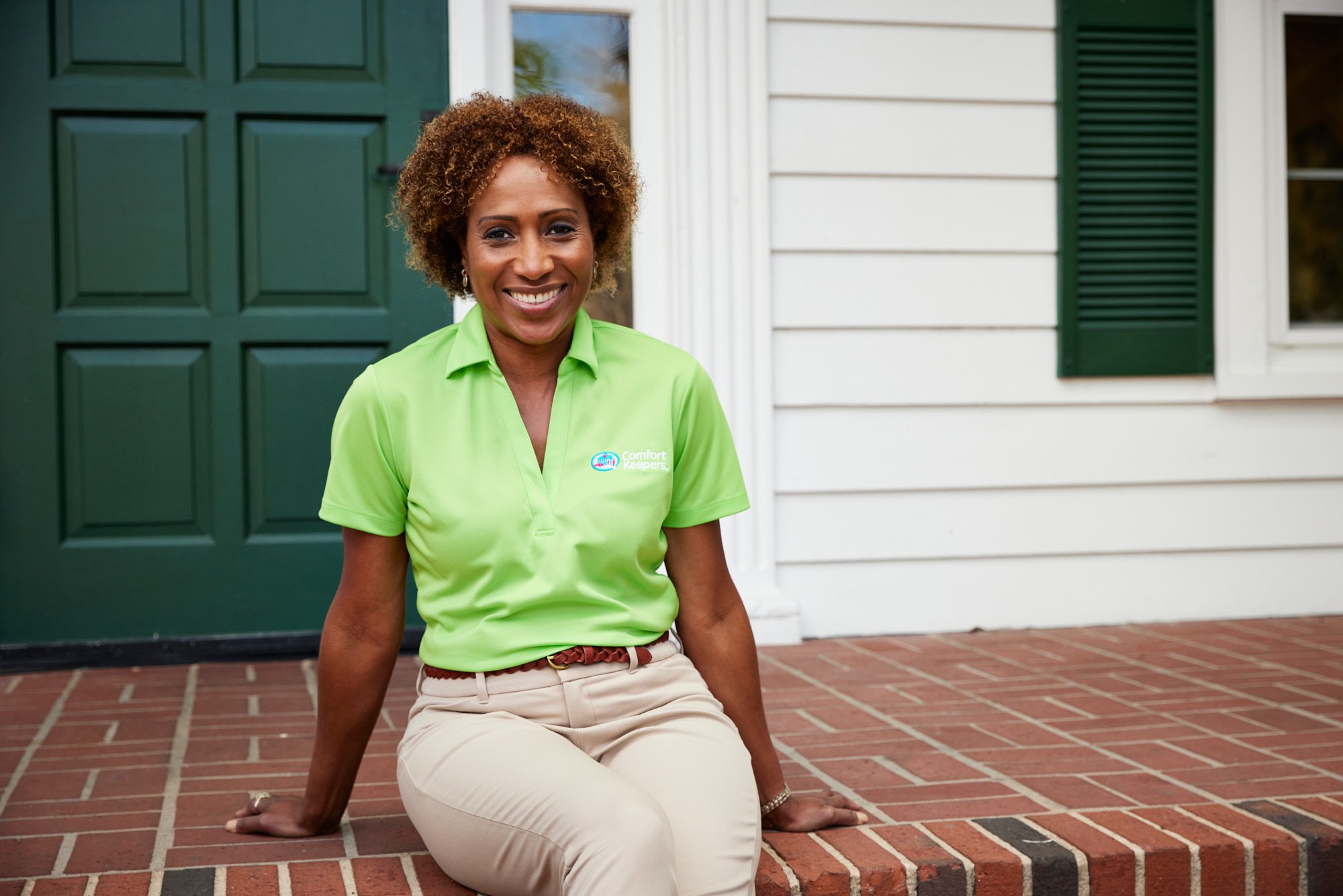 Smiling person in green polo with "Comfort Keepers" logo sitting on brick steps in front of green door.