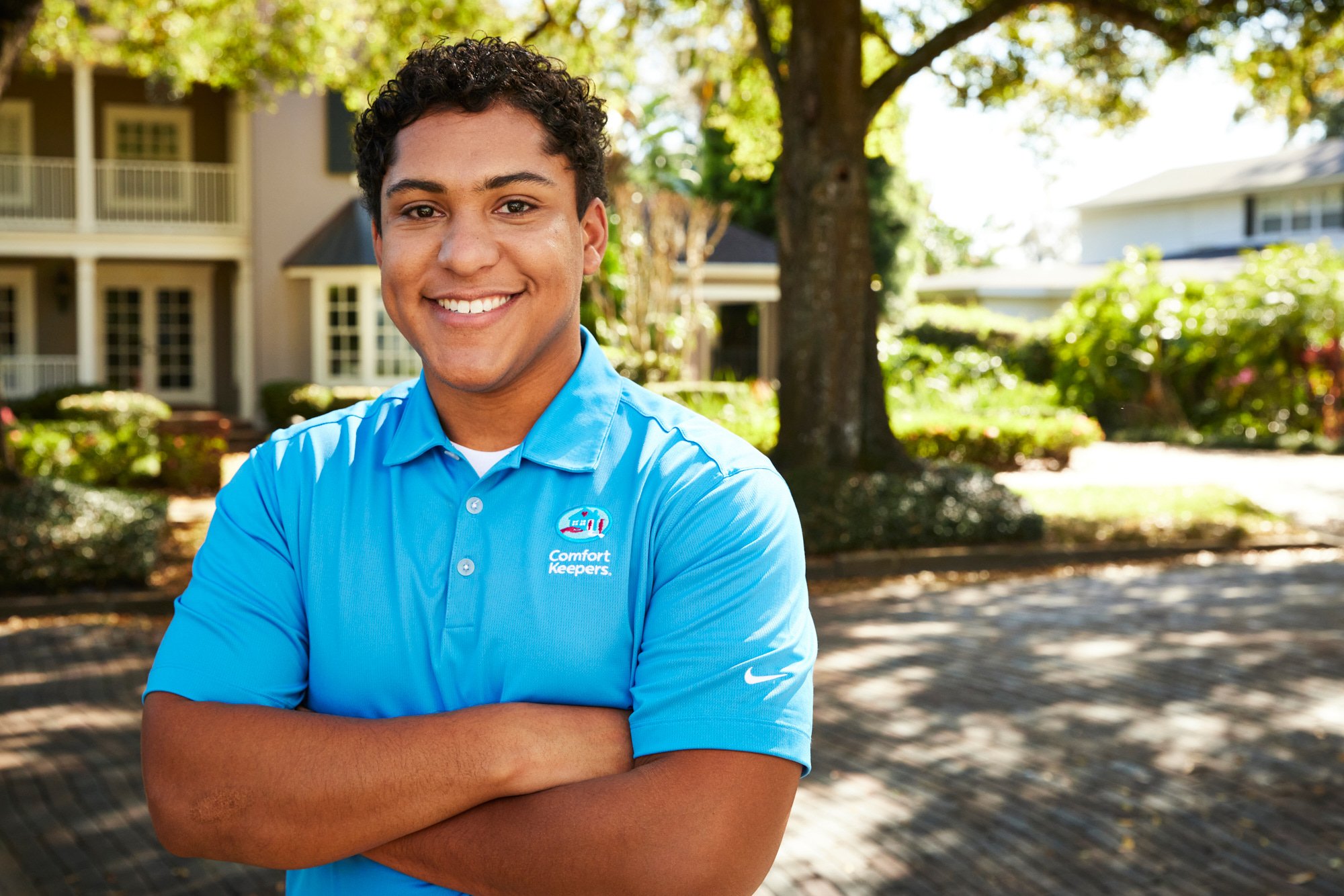 Smiling person in blue Comfort Keepers polo with crossed arms, standing outdoors in a shaded area near a building.
