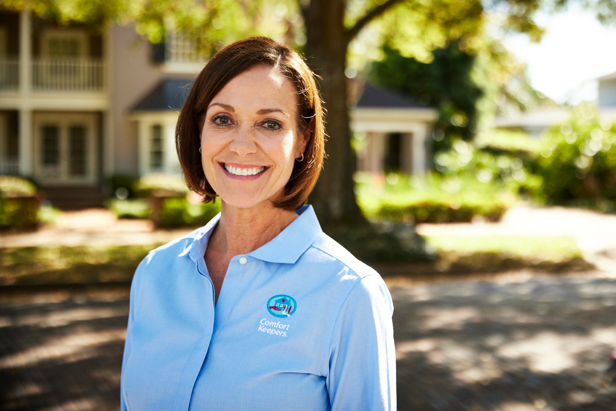 Smiling woman in blue Comfort Keepers shirt outside a house with trees in the background.