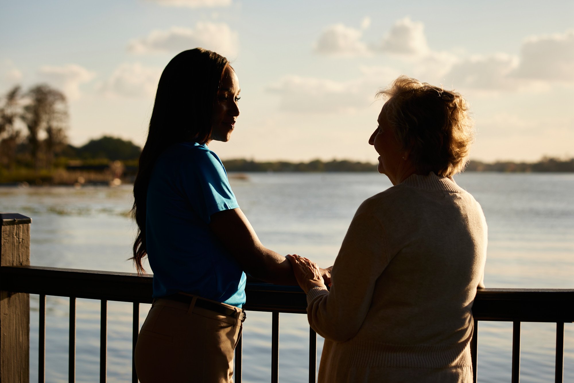 Two women standing near a railing, holding hands, with a scenic lakeside view in the background.