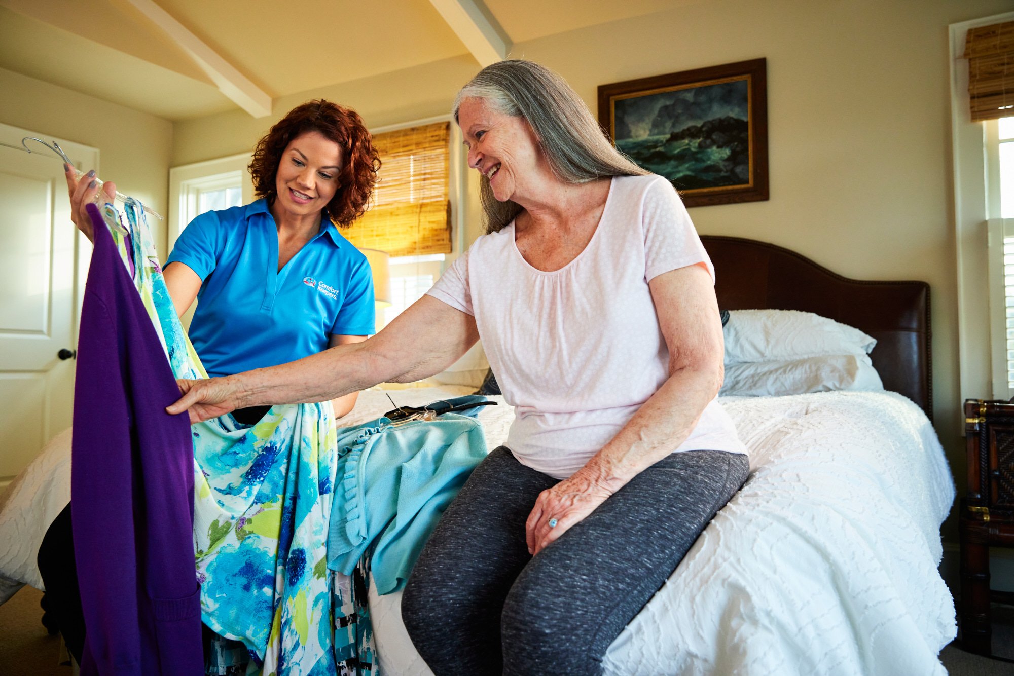 A caregiver in a blue shirt assists an elderly woman sitting on a bed with selecting clothes.