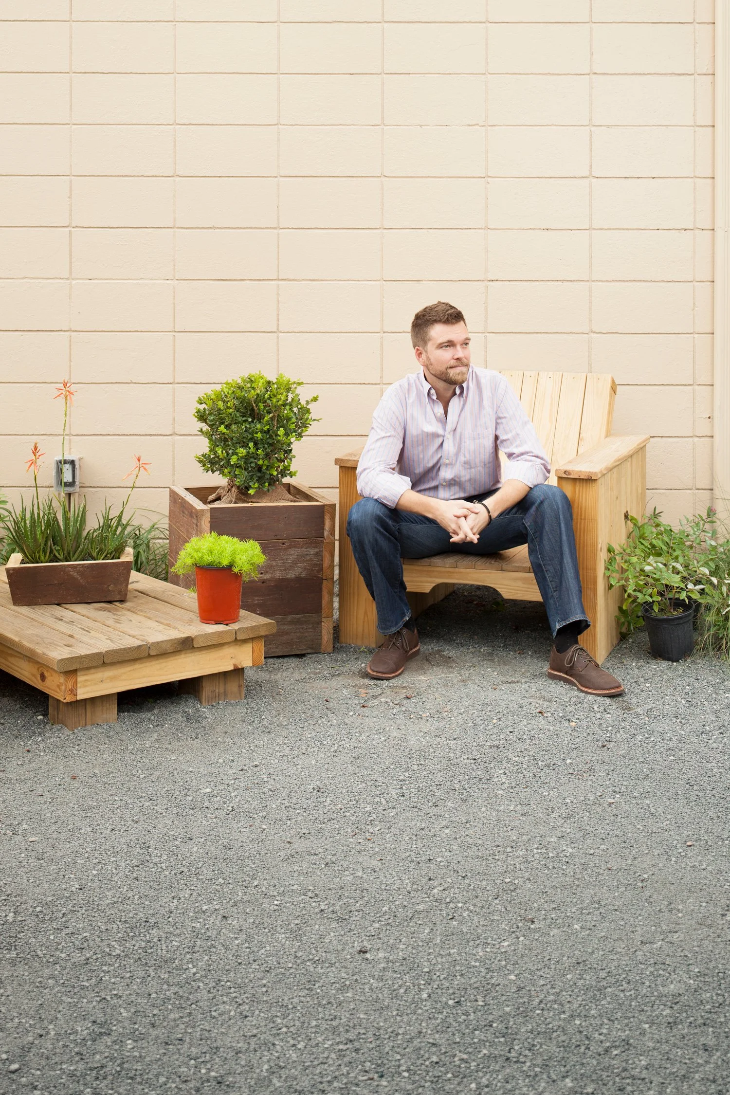 A man with short brown hair and a beard sitting on a wooden bench outdoors near a beige brick wall, surrounded by potted plants.