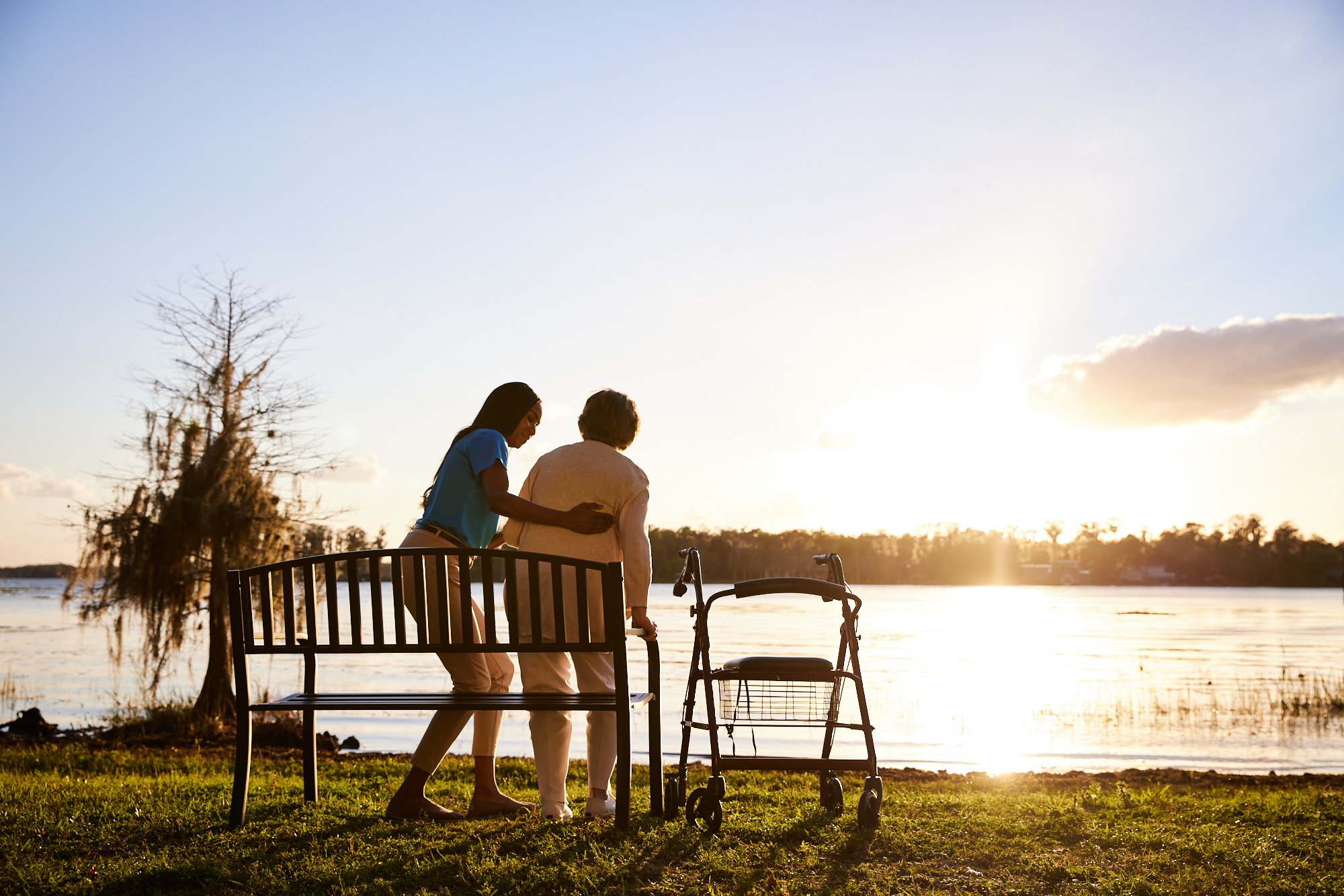 A caregiver assisting an elderly person near a lake at sunset, with a walker nearby and both standing beside a bench.