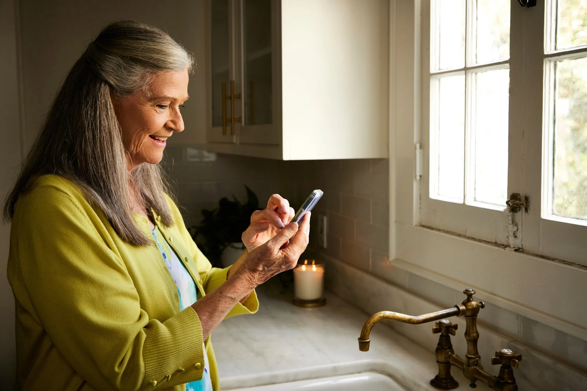 Older woman using a smartphone in a kitchen with a lit candle and window.