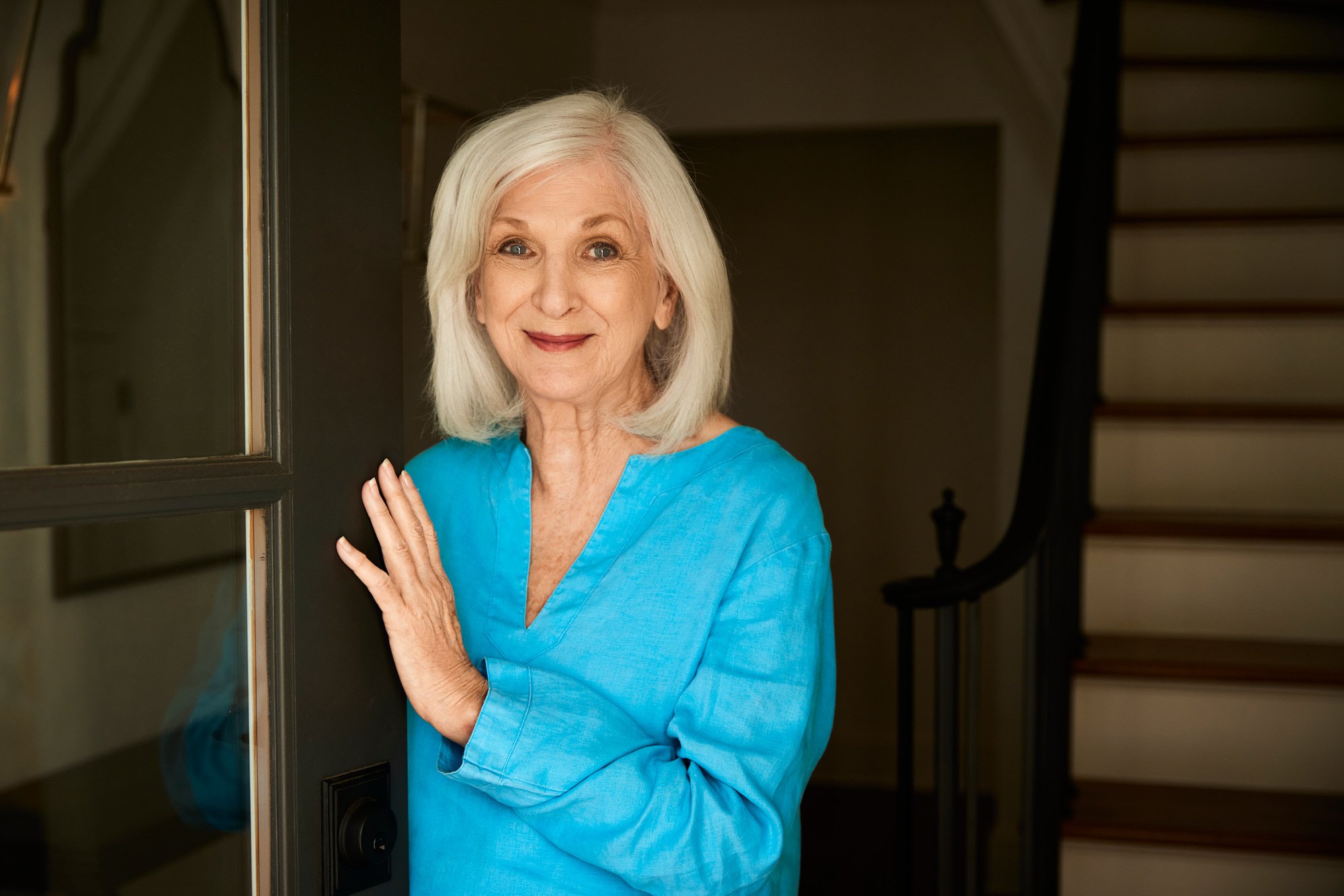 Elderly woman with white hair wearing a blue shirt standing by a doorway and smiling.