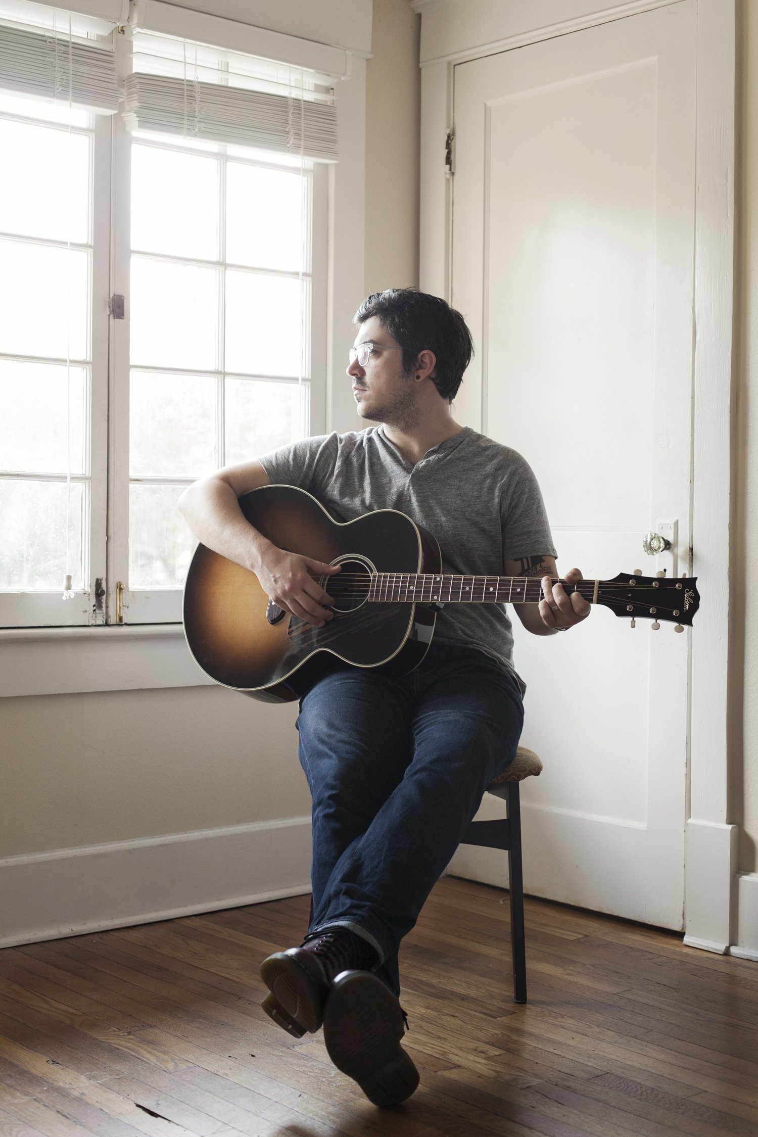 A man with glasses and dark hair sits on a chair playing an acoustic guitar in a room with large windows and hardwood floors.