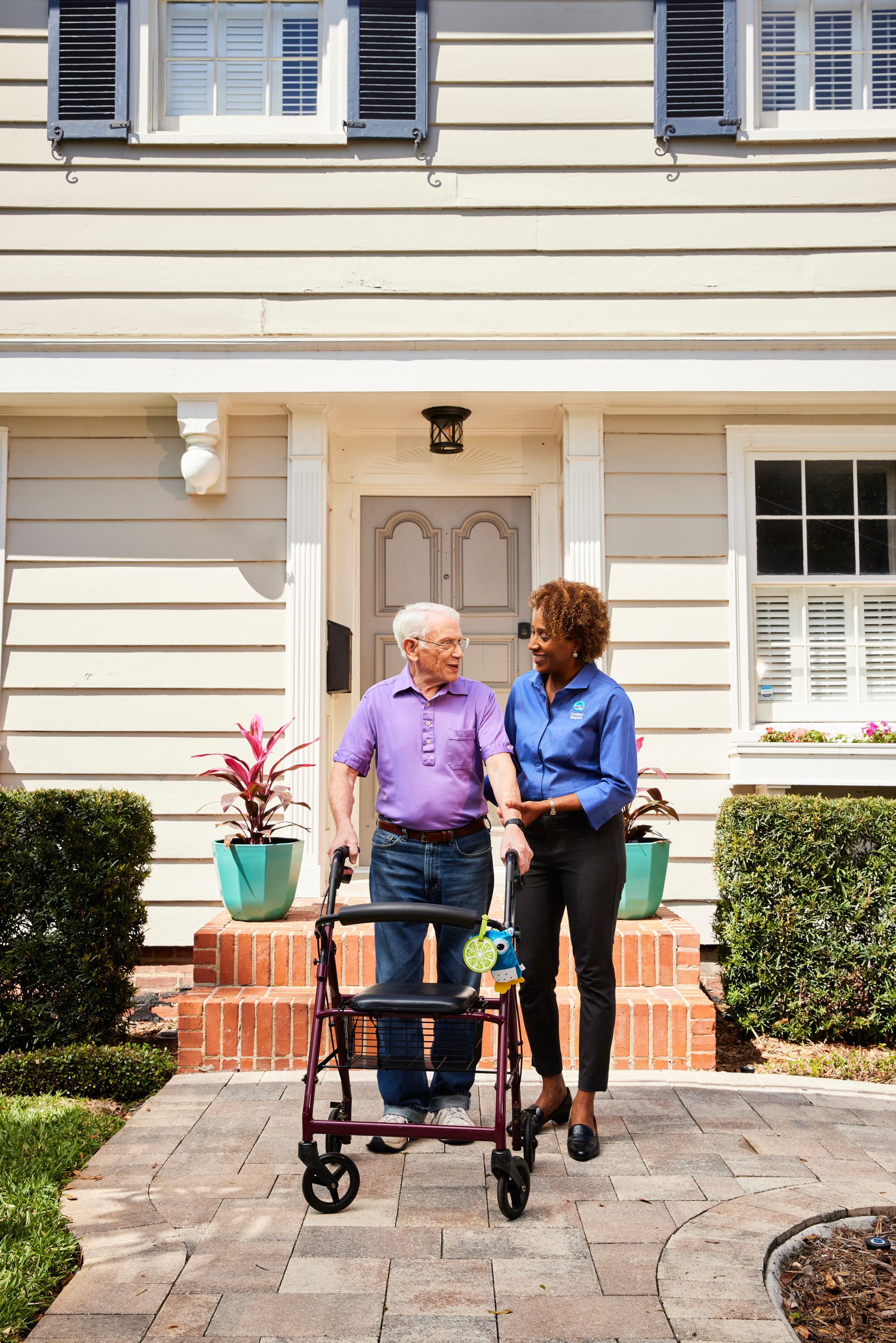 A caregiver assisting an elderly man with a walker in front of a house.
