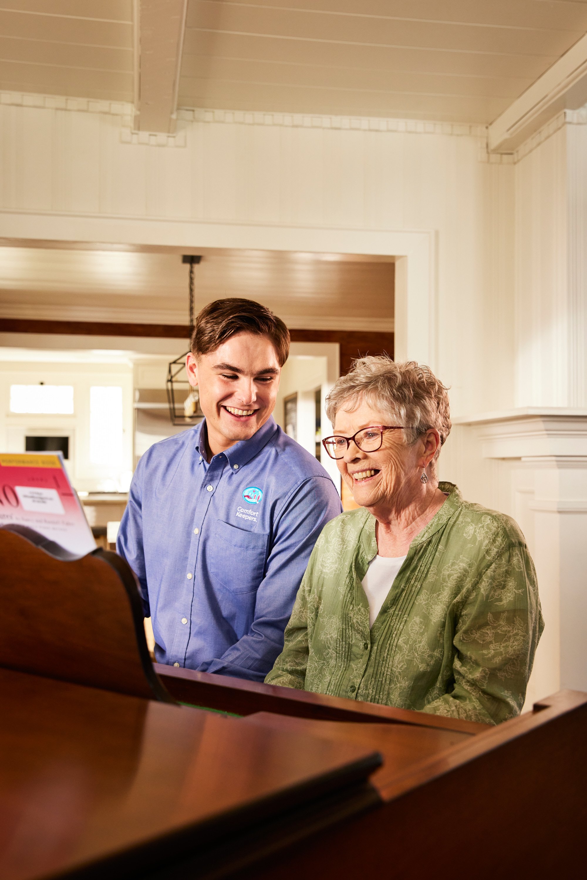 A young man and an elderly woman smiling and sitting at a piano in a warmly lit room.