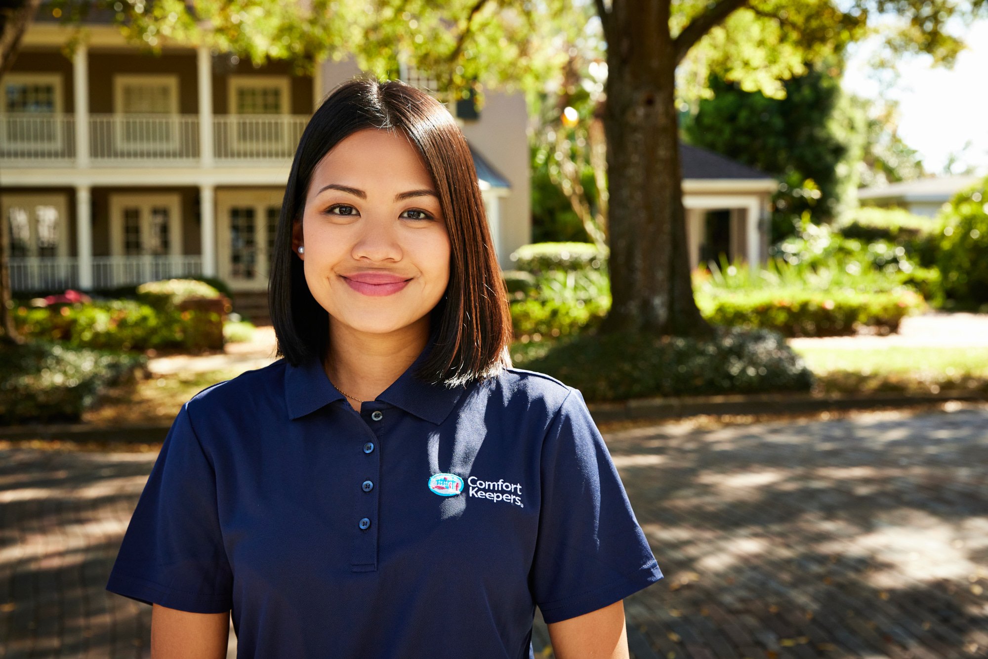 Smiling woman wearing a navy blue "Comfort Keepers" polo shirt, standing outdoors with trees and a building in the background.