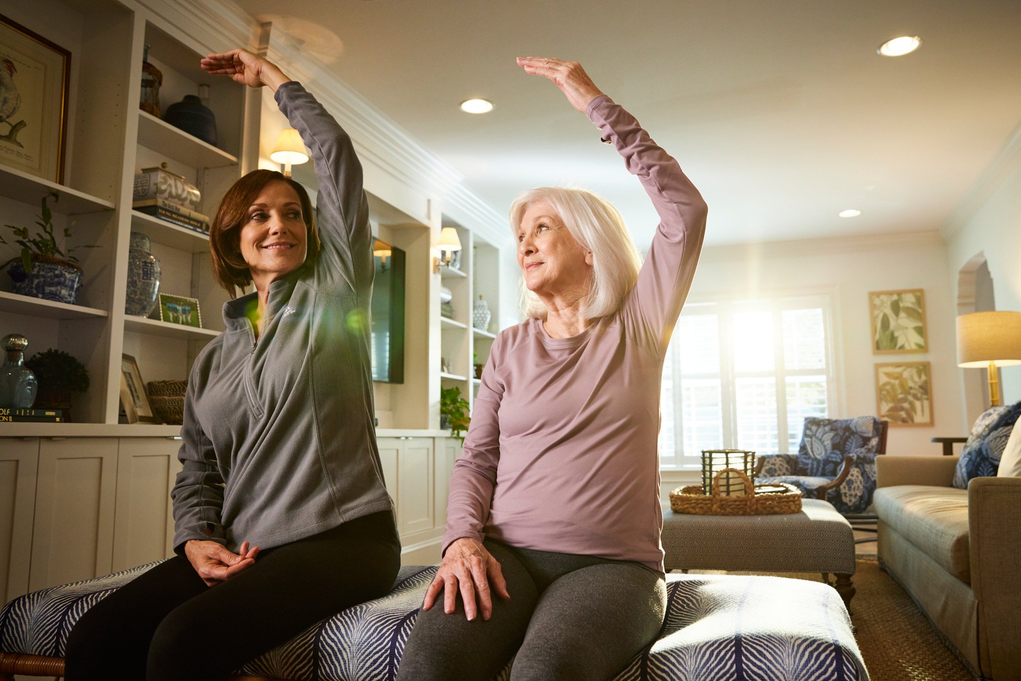 Two women sitting indoors, stretching with one arm raised, in a well-lit living room with comfortable furniture and shelves in the background.