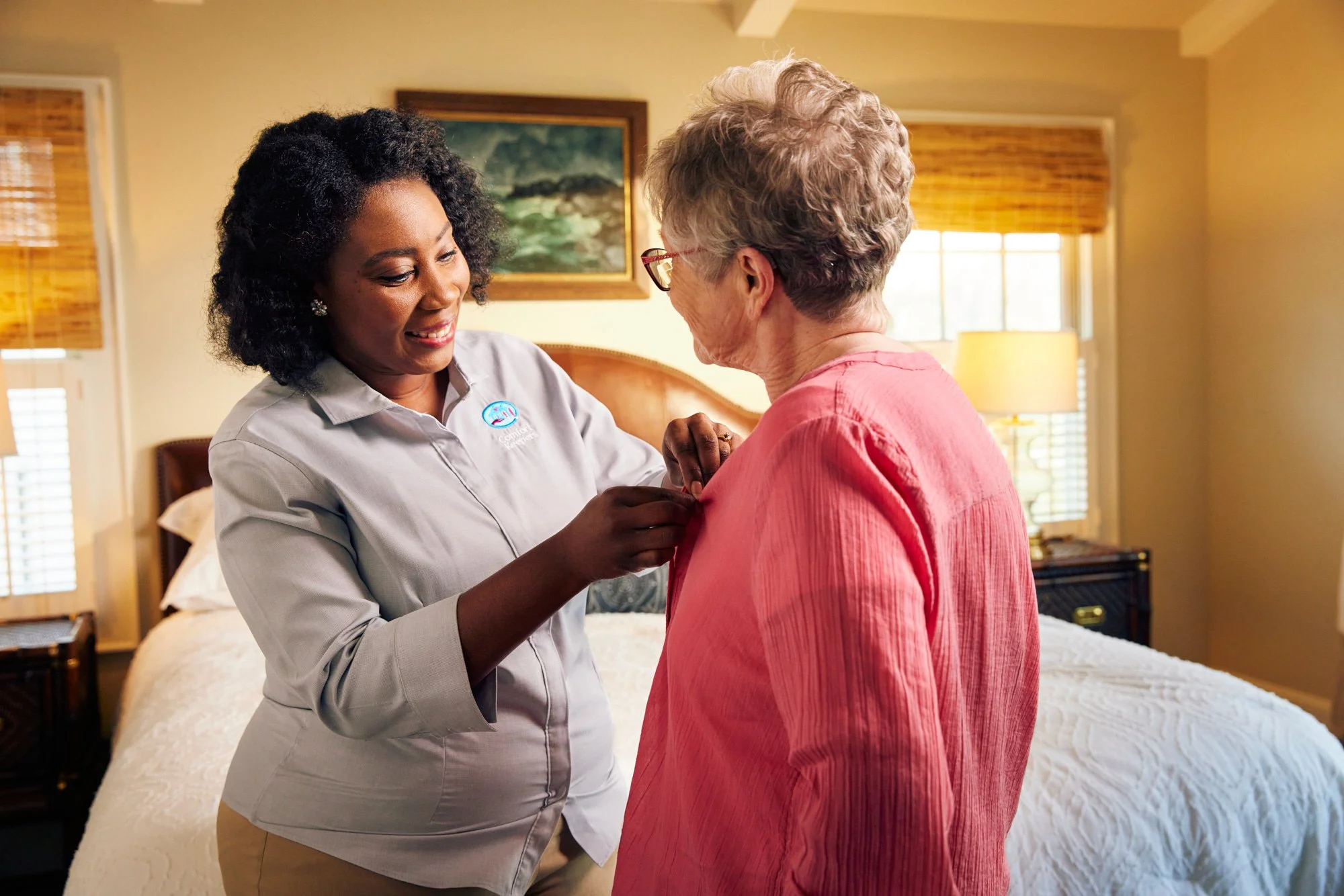 A caregiver helping an elderly woman button her shirt in a bedroom setting. The caregiver is smiling and wearing a uniform with a logo. The elderly woman is wearing a pink shirt and has glasses.