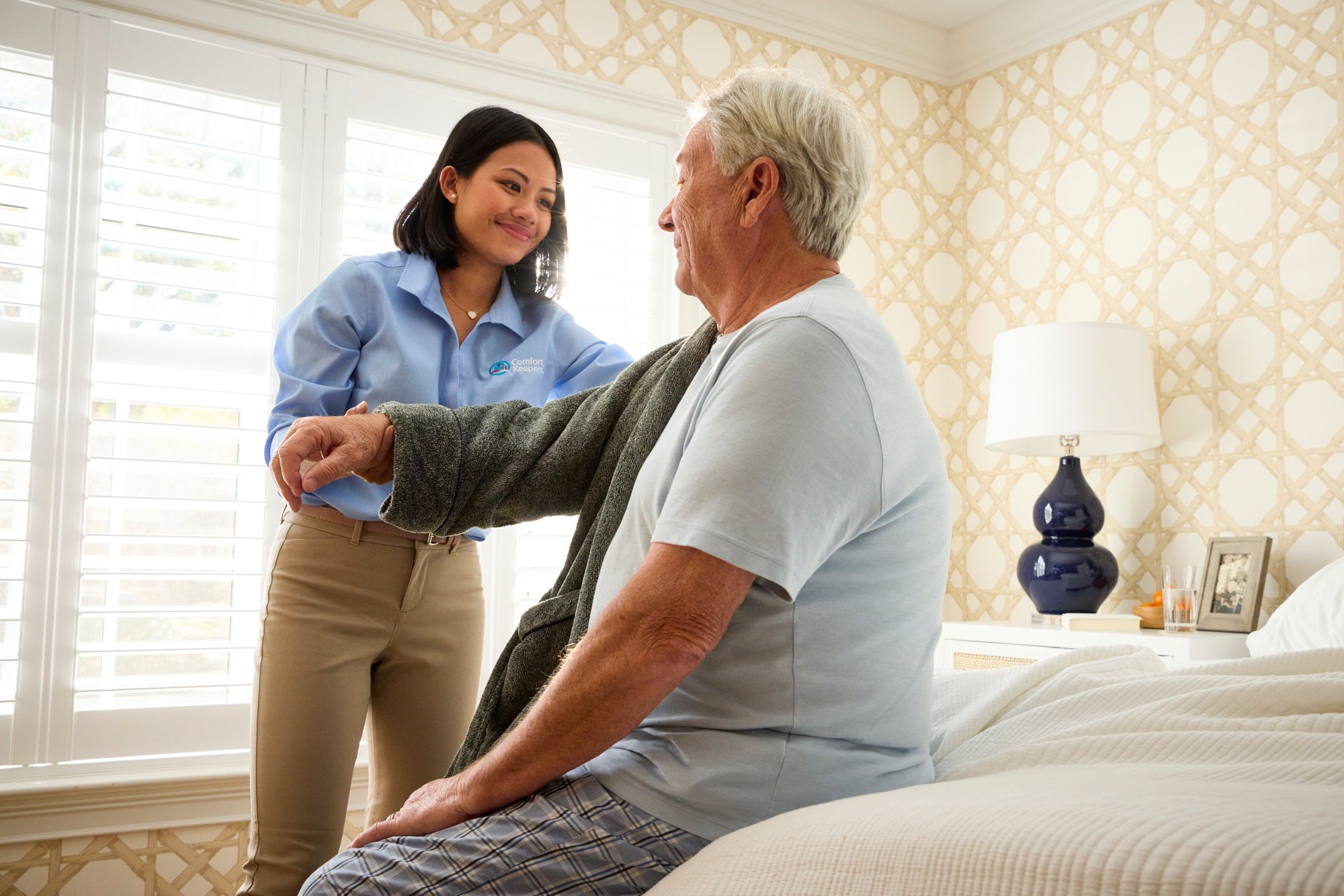 A caregiver assisting an elderly man in a bedroom, helping him with his robe.