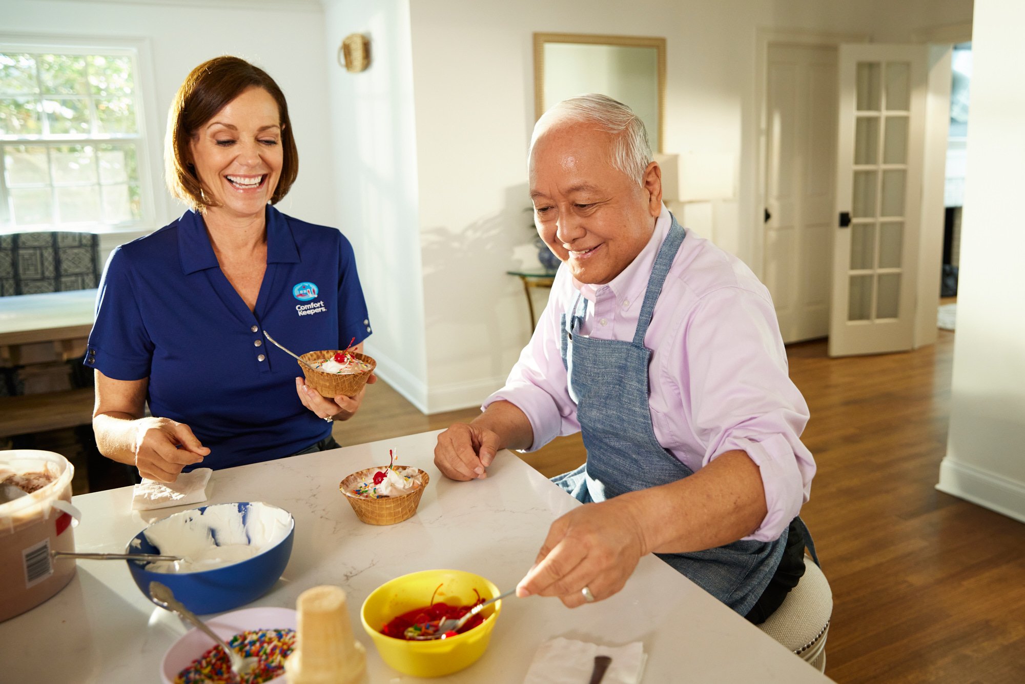 Two people making ice cream sundaes in a kitchen with toppings and waffle bowls.