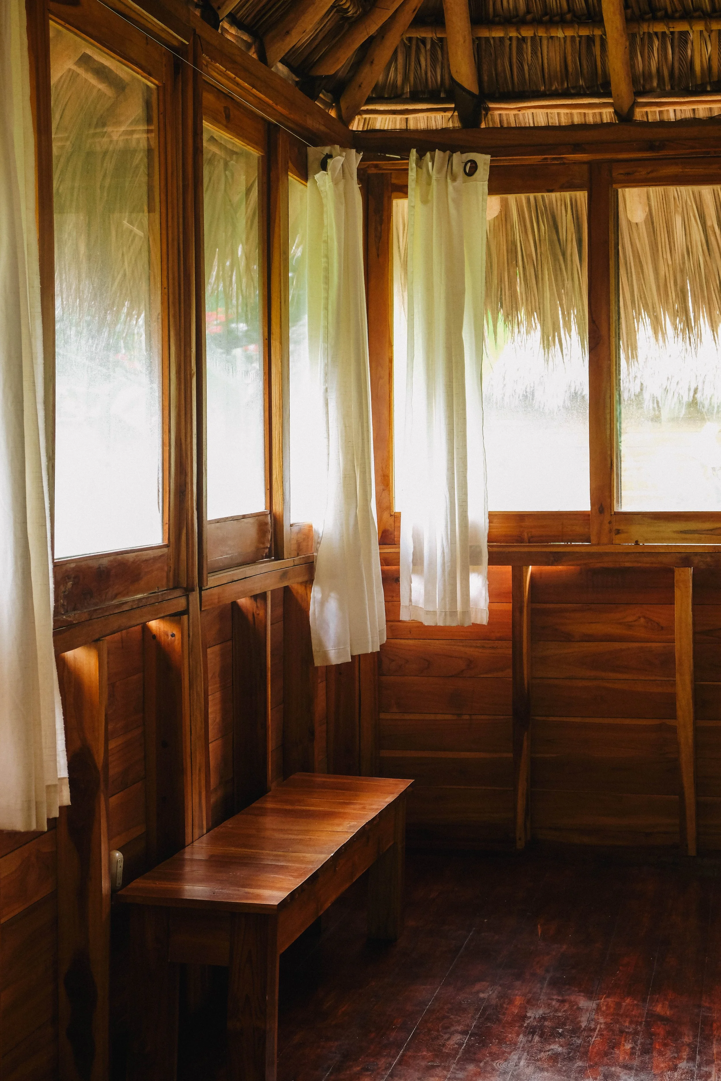 Wood-paneled upstairs cabaña room with sheer-curtained windows and a thatched roof at Still Salty Escape surf and yoga retreat in Nicaragua